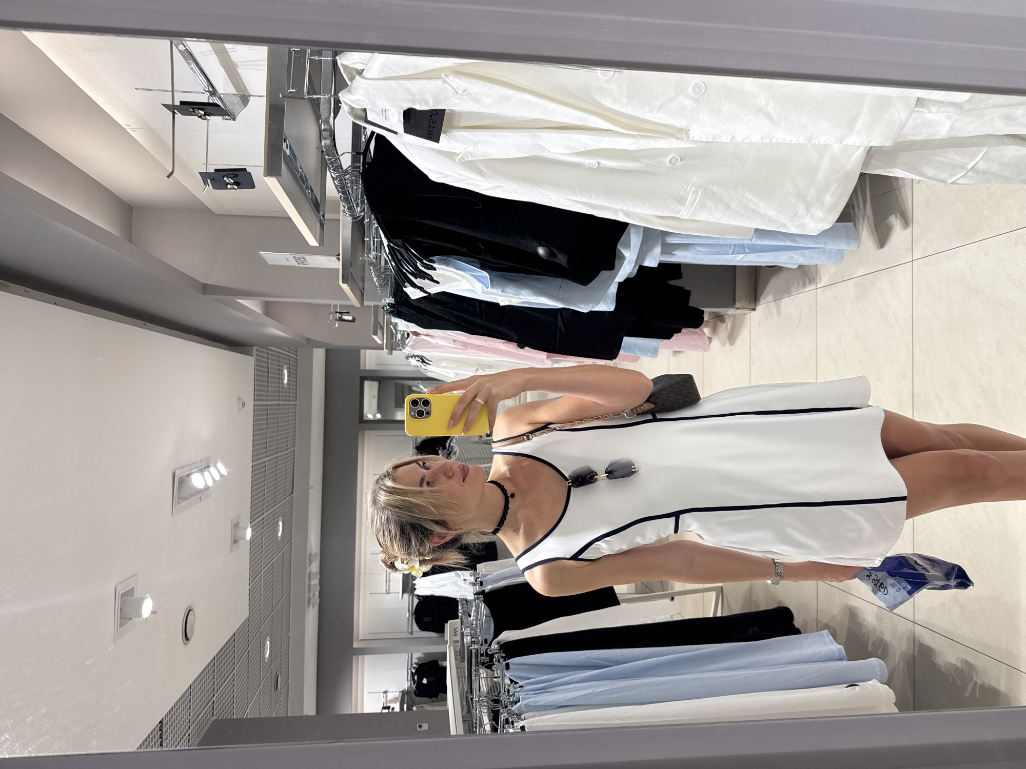 A mirror-reflected view shows a woman taking a selfie in a clothing store in an unknown city. She is holding a yellow smartphone in her left hand and a blue shopping bag in her right hand. She is wearing a white dress with black trim and has sunglasses hooked on the dress. The woman also has a black choker necklace. Clothes on hangers are seen on racks in the store. The floor is tiled.