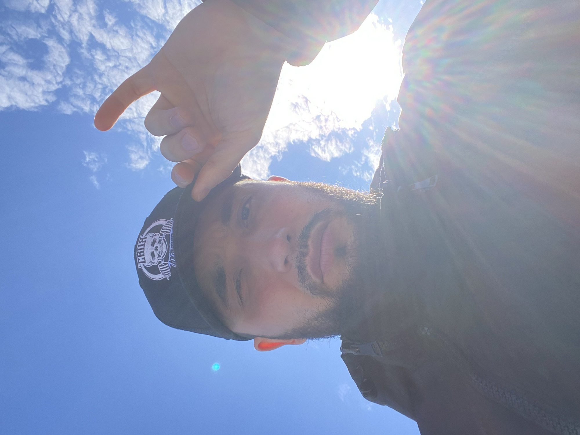 A low-angle, selfie-style photograph shows a man with medium skin tone, a dark beard, and a black baseball cap with a white skull and barbell logo. He is partially backlit by the sun, causing lens flare. He is pointing with his left index finger towards the blue sky with scattered white clouds. The man is wearing a dark jacket. The location is Koskolovo, Russia.