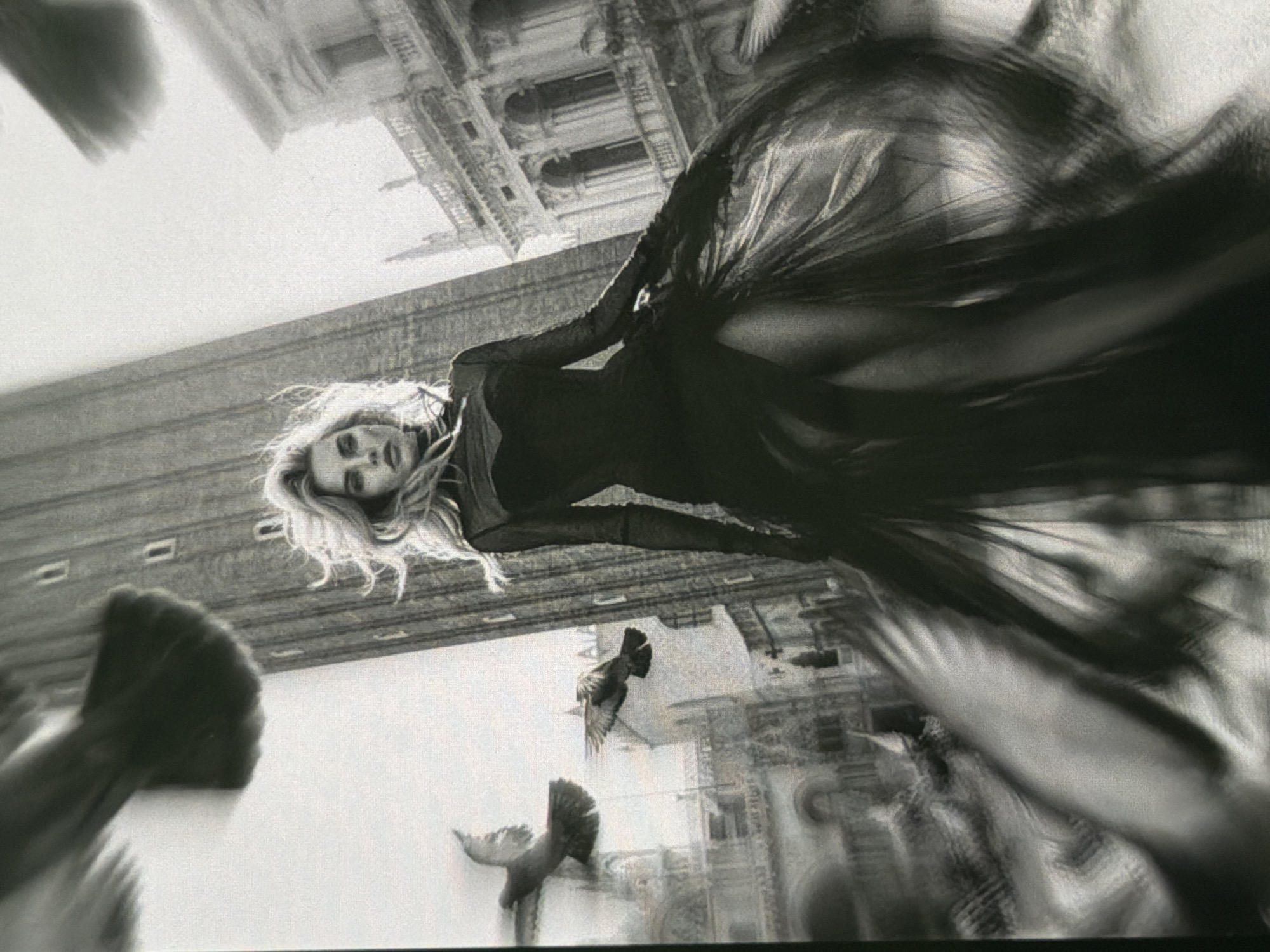 A black and white, slightly blurred image depicts a woman in a flowing, dark gown standing in what appears to be a city square. She has blonde, wind-swept hair and is looking towards the camera. In the background, a tall, slender tower dominates the skyline, with ornate buildings flanking it. Several pigeons are captured in mid-flight around the woman, adding a sense of movement to the scene. The architecture suggests a European setting, possibly Venice, Italy, with its canals and historical buildings. There is no visual information to suggest this location is Bobrovo, Russia. The image captures a dramatic and artistic moment.