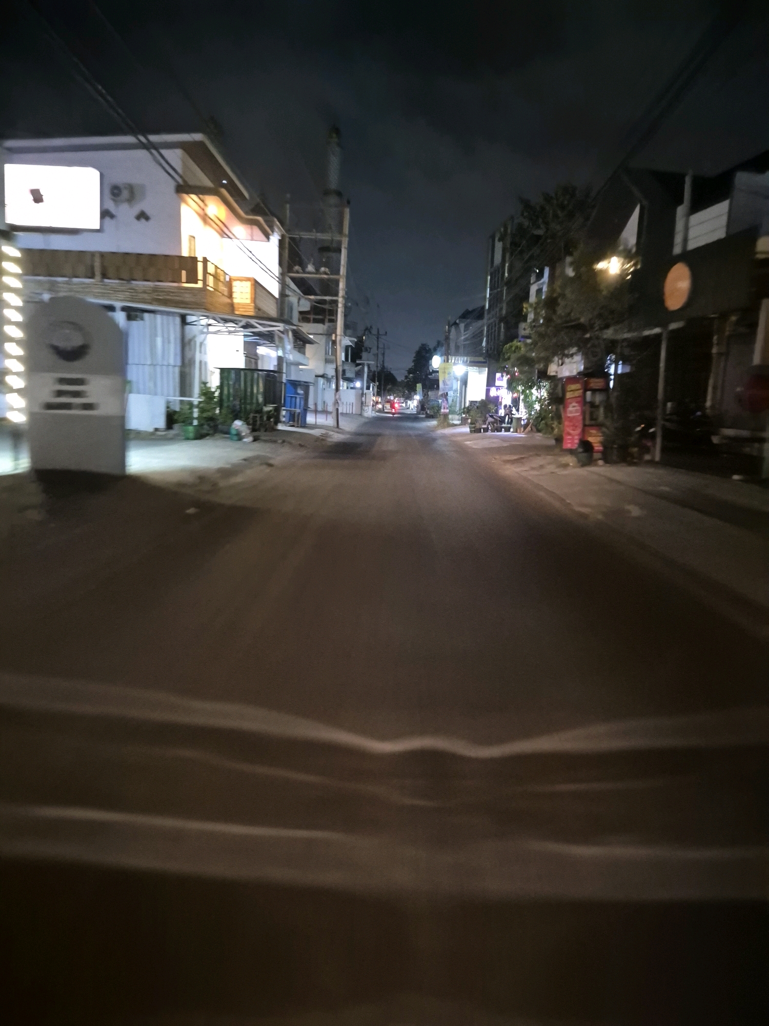 The image depicts a street scene at night in Pabelan, Indonesia. The street is paved and illuminated by streetlights and lights from the surrounding buildings. On the left side of the street, there is a two-story building with a white facade and warm-toned lighting on the upper floor. A large, illuminated sign is visible on the left side of this building. In front of the building, there is a grey, pillar-like monument with some text and a circular emblem. Further down the street, there are more buildings, some of which appear to be shops or restaurants, with glowing signs and lights. There are also some trees and greenery visible. The street appears to be relatively quiet, with no people or vehicles prominently featured in the foreground. The sky is dark, indicating nighttime.