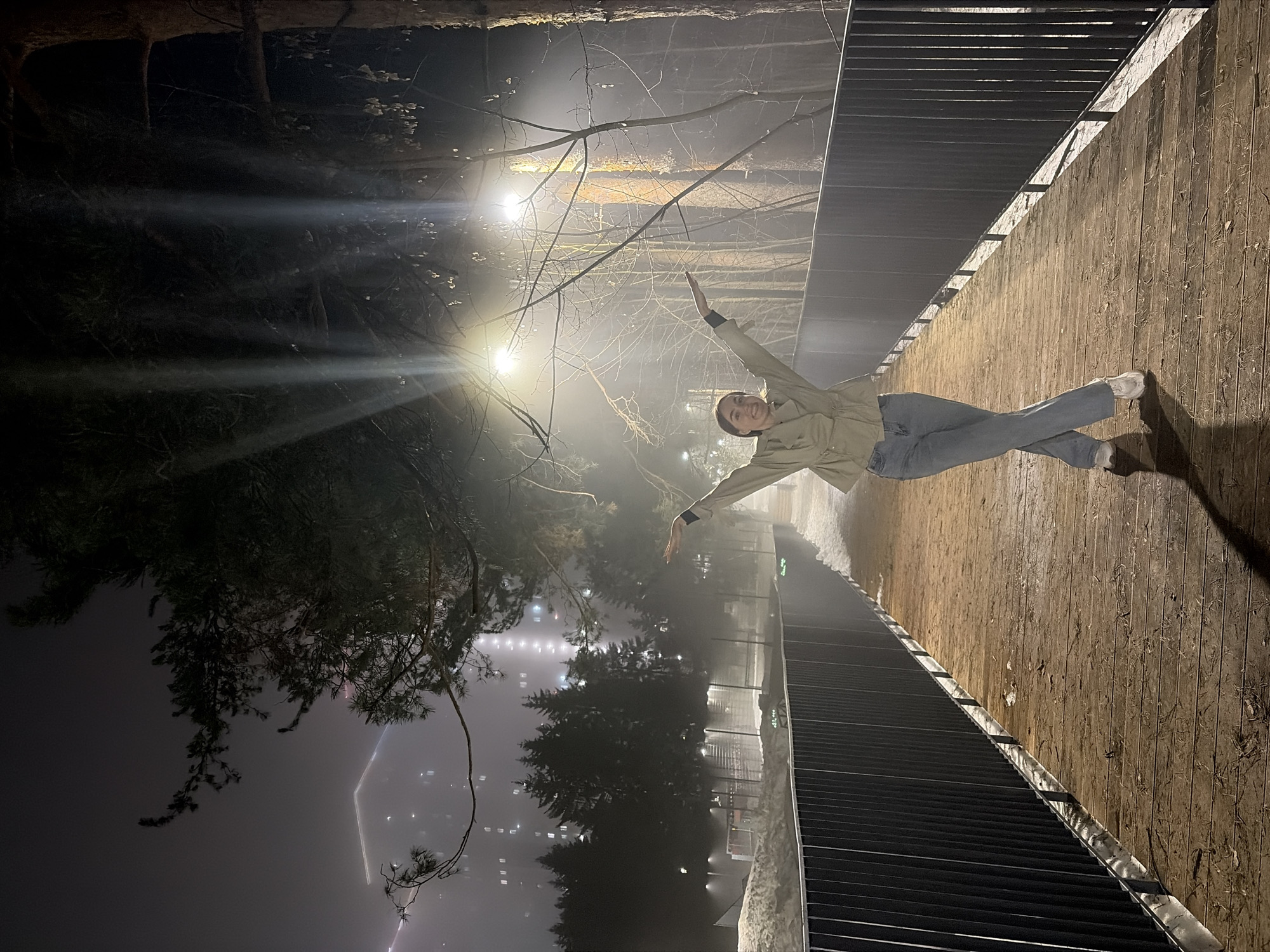 A woman stands on a wooden bridge, posing with her arms outstretched in a foggy night environment in Izhevsk, Russia.
The woman is wearing a light-colored jacket, blue jeans, and white shoes. She is smiling.
The bridge is made of wood and has metal railings. The surrounding environment is a foggy park with trees and lights. In the background, city lights and a tall building can be seen through the fog. The scene is illuminated by streetlights.