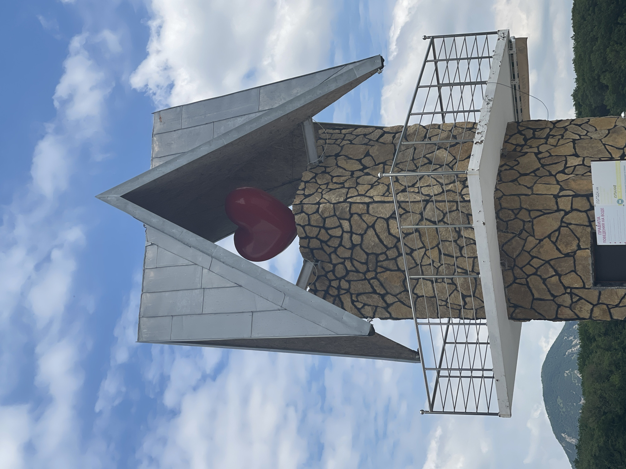 The image shows a stone monument topped with a large red heart sculpture. The monument has a metal roof that forms a pointed arch. A white platform with a metal railing encircles the upper portion of the monument. In the background, there are green trees and a mountain. The sky is blue with scattered white clouds. Text is visible on a sign at the base of the monument, but it is not legible. This monument is located in the city of Zheleznovodsk, Russia.