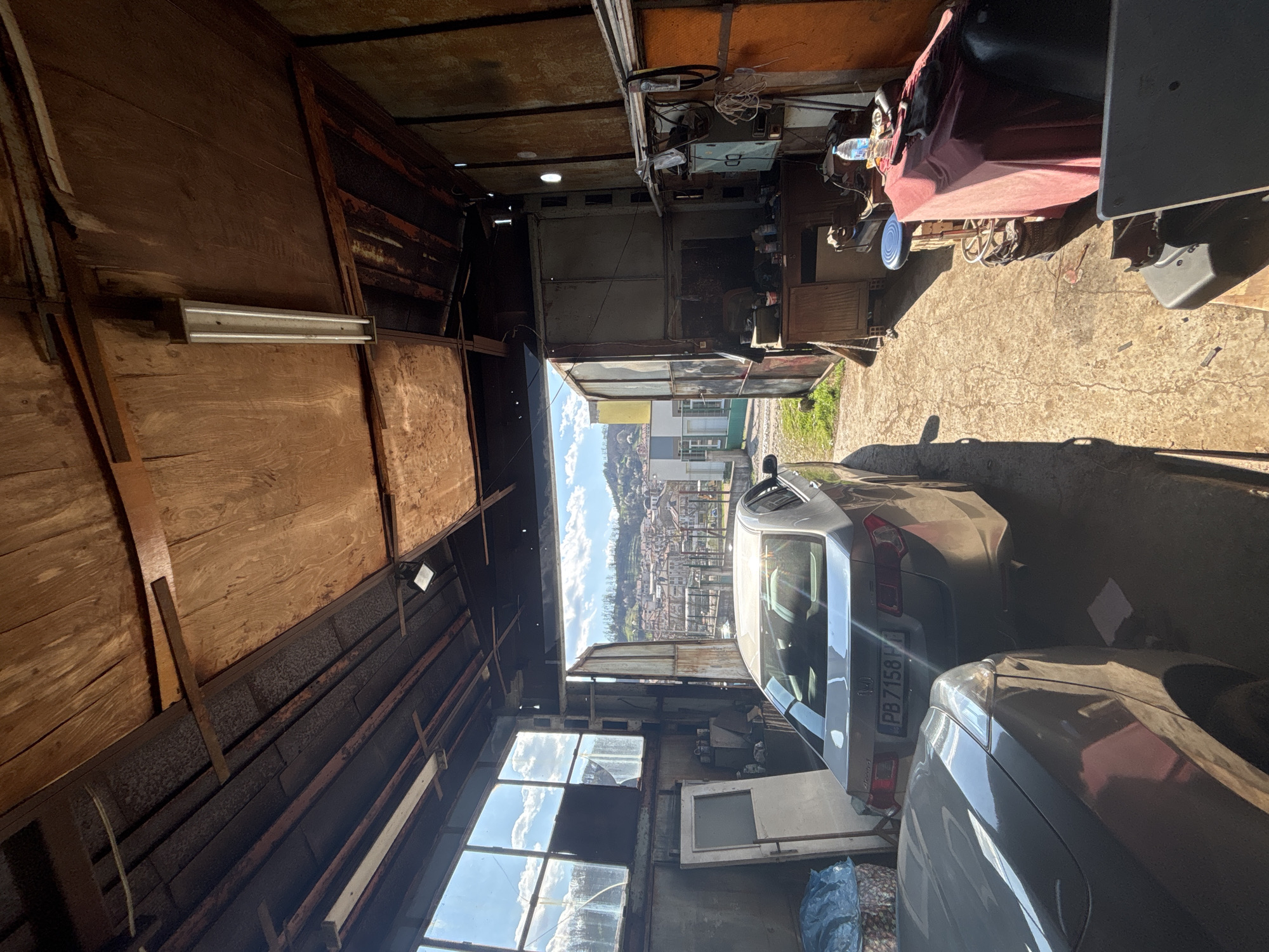 The image presents an interior view of a cluttered garage in Zlatograd, Bulgaria, looking out towards the city. The garage is constructed from wood and metal, showing signs of age and disrepair. Several vehicles are parked inside, partially obscuring the view towards the cityscape. Natural light illuminates the interior, highlighting the urban landscape visible beyond the garage opening. The scene appears to be a residential area.