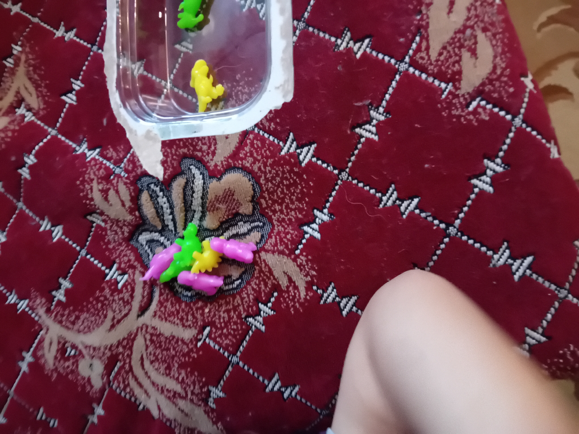An overhead shot of toys on a red carpet. In the center of the image are several small plastic toys, one green, one yellow, and two pink, arranged in a cluster. To the right, a clear plastic container holds a yellow and a green plastic dinosaur toy in water. The carpet has a diamond pattern with light-colored stitching. In the lower left corner, a person's bare leg is visible. The location is not specified.