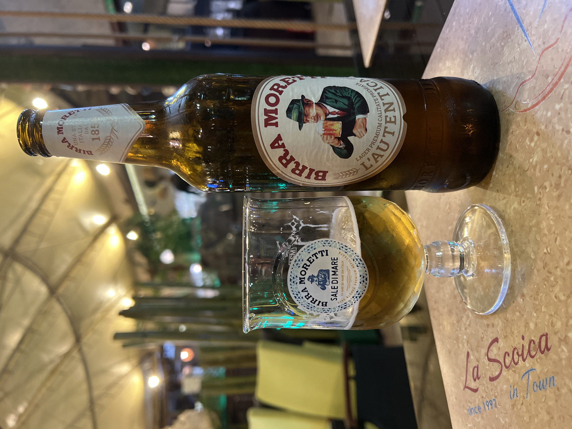 An amber glass bottle of Birra Moretti beer and a glass filled with the same beer are on a table. The bottle is standing upright on the right side of the image, and the glass is on the left. The labels on both the bottle and glass are visible. The background shows a blurry outdoor setting with lights and some greenery. The table has a light brown patterned surface with some faded red and blue lines and text that says "La Soica in Town since 1997". The overall lighting suggests it is nighttime or a dimly lit indoor space. The presence of the text "La Soica in Town since 1997" strongly suggests the location is La Soica restaurant in Constanța, Romania.