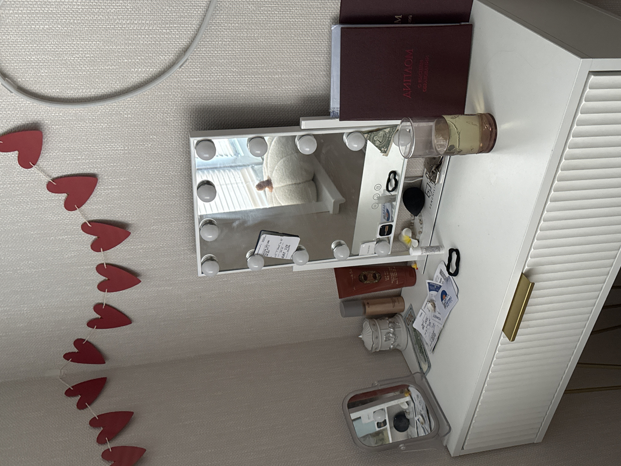 The image shows a vanity dresser with various items on its surface. The dresser is white with a gold handle and ribbed detailing on the drawer front. On the dresser, there is a lighted makeup mirror, a small decorative jar, a bottle of lotion, a red tube of cream, and a glass of yellowish liquid. Several papers and tickets are scattered on the surface. To the right of the mirror are two burgundy-colored diplomas, one with the word "ДИПЛОМ" visible in red lettering. In the background, a string of red paper hearts is hung on a textured wall. The overall scene suggests a personal vanity area. There is no indication of the location being in the city of Perm, Russia.