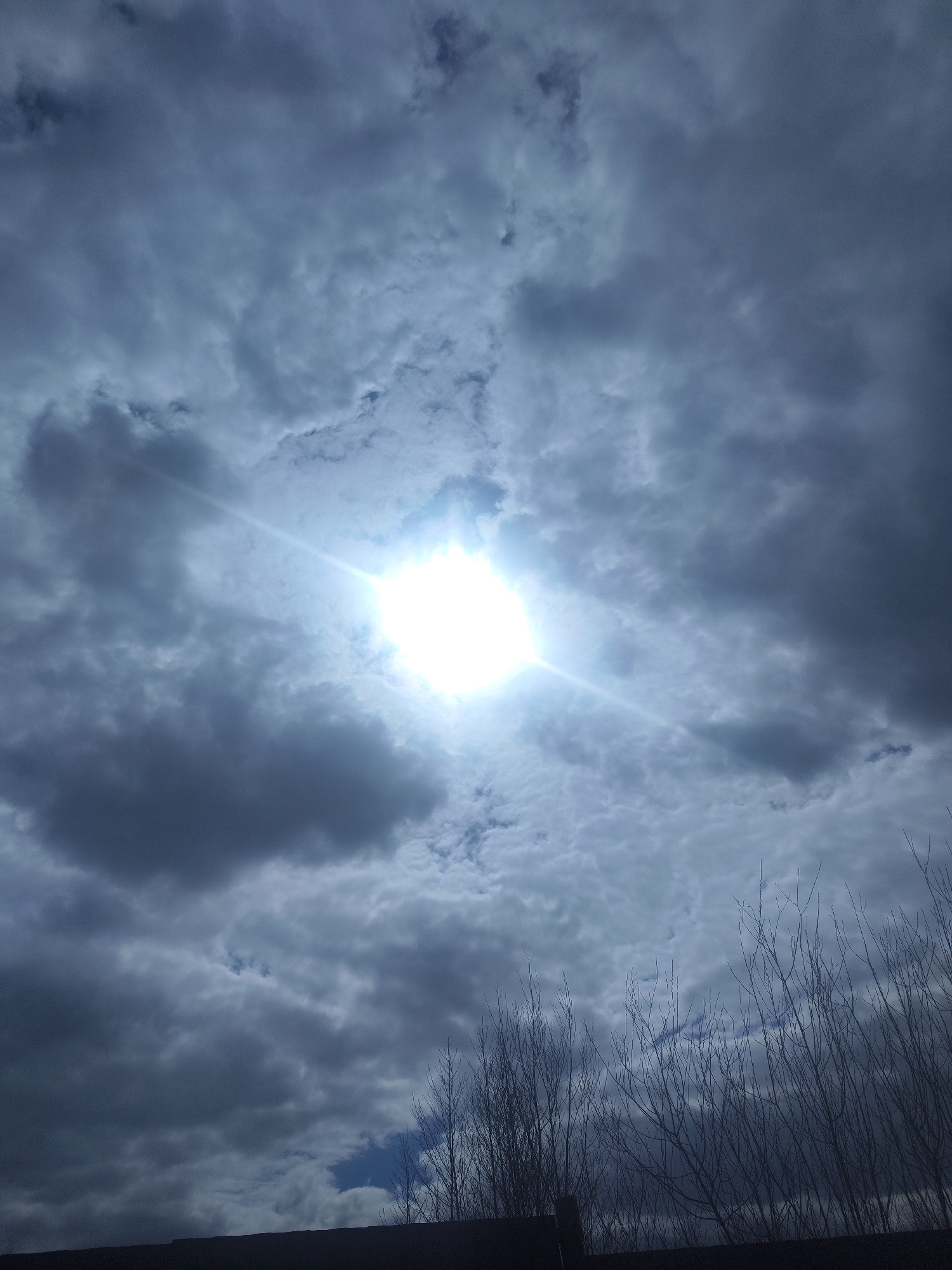The image shows a sky with dense, dark gray clouds. A bright sun is visible through a break in the clouds, creating a lens flare effect. In the foreground, the silhouettes of leafless tree branches are visible. The location is Unknown, Unknown. There are no people in the image.