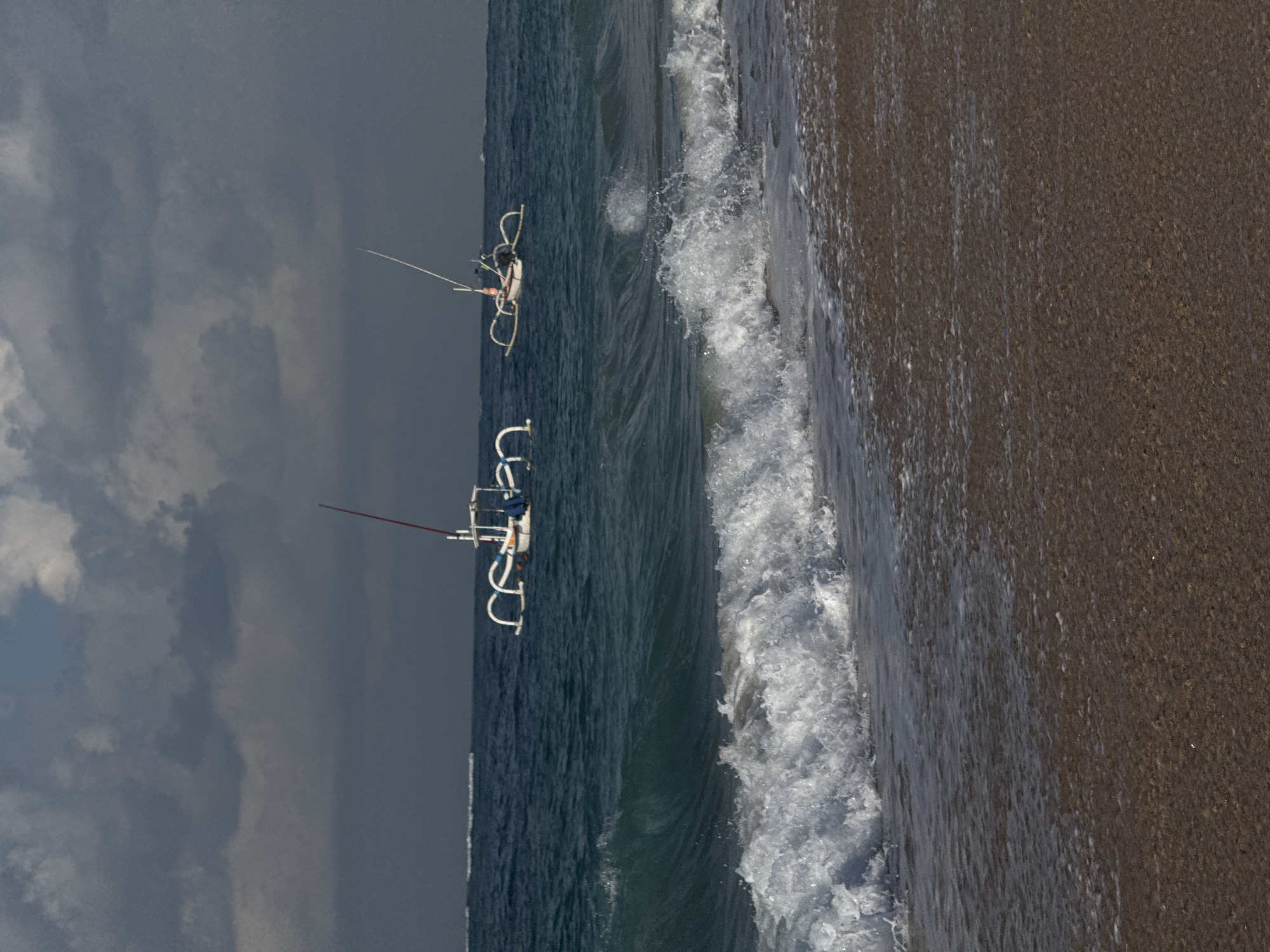 The image depicts a coastal scene in Kuta Selatan, Indonesia. The foreground features a sandy beach with gentle waves lapping the shore, creating white foam. In the middle ground, the turquoise ocean stretches out, with a series of white-capped waves rolling in. Two traditional outrigger boats, commonly known as jukung, are visible on the water. These boats are white with distinctive outrigger poles extending outwards. One boat is closer to the viewer and has a red pole extending upwards. The other boat is further in the distance. The background is dominated by a dark, cloudy sky, suggesting overcast weather. The horizon line is clearly defined between the sea and the sky.