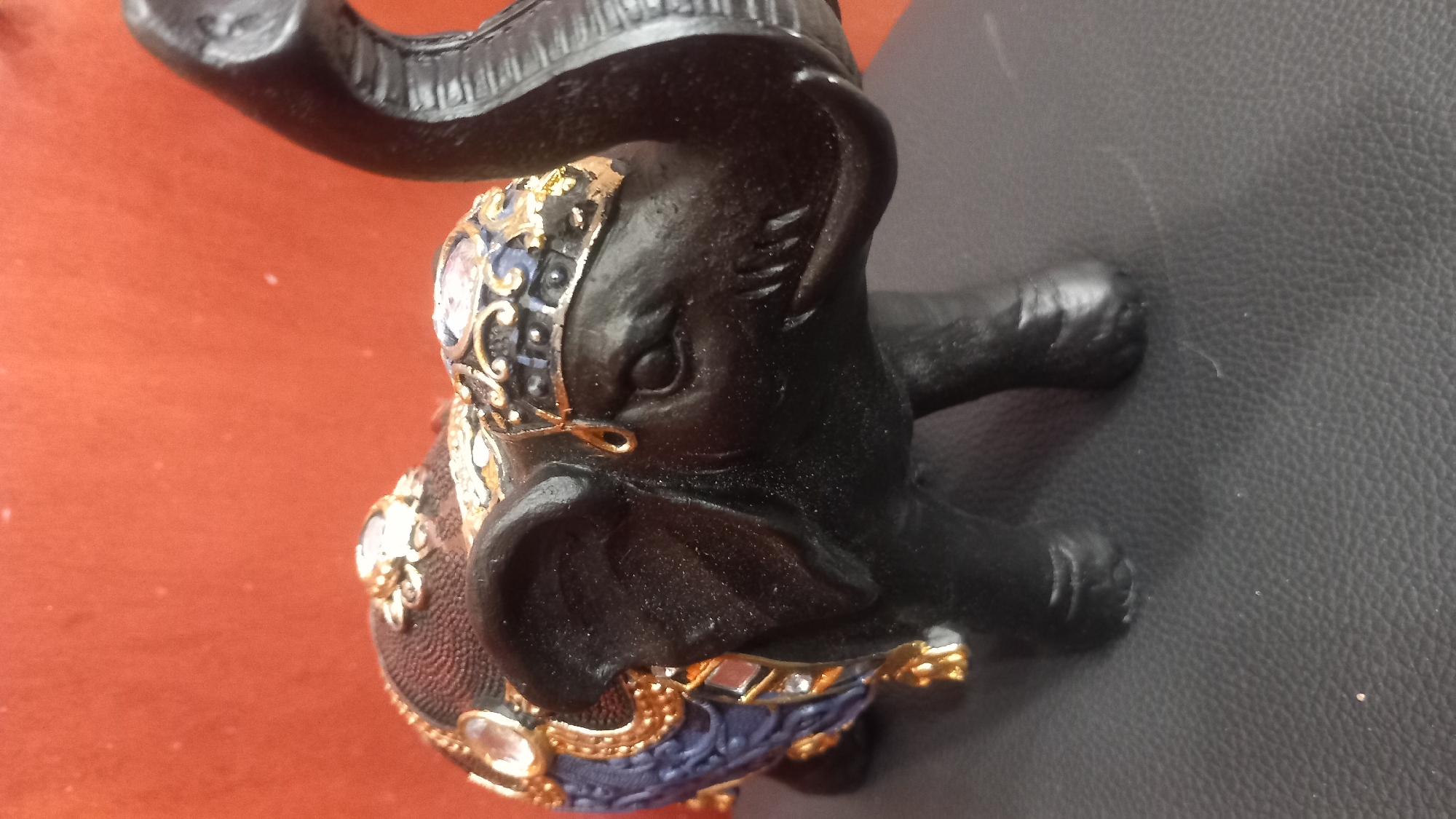 A black, decorative elephant figurine is positioned on a dark, textured surface, likely a leatherette material. The elephant is adorned with elaborate gold trim, blue fabric accents, and clear, faceted embellishments that appear to be rhinestones or similar faux gems. The background is a solid, warm orange-red color. The figurine's trunk is raised, and its mouth is slightly open, revealing small, pointed tusks. The elephant's ears are large and prominent. There is no indication of the city of Kos, Greece, or any urban environment within the image.