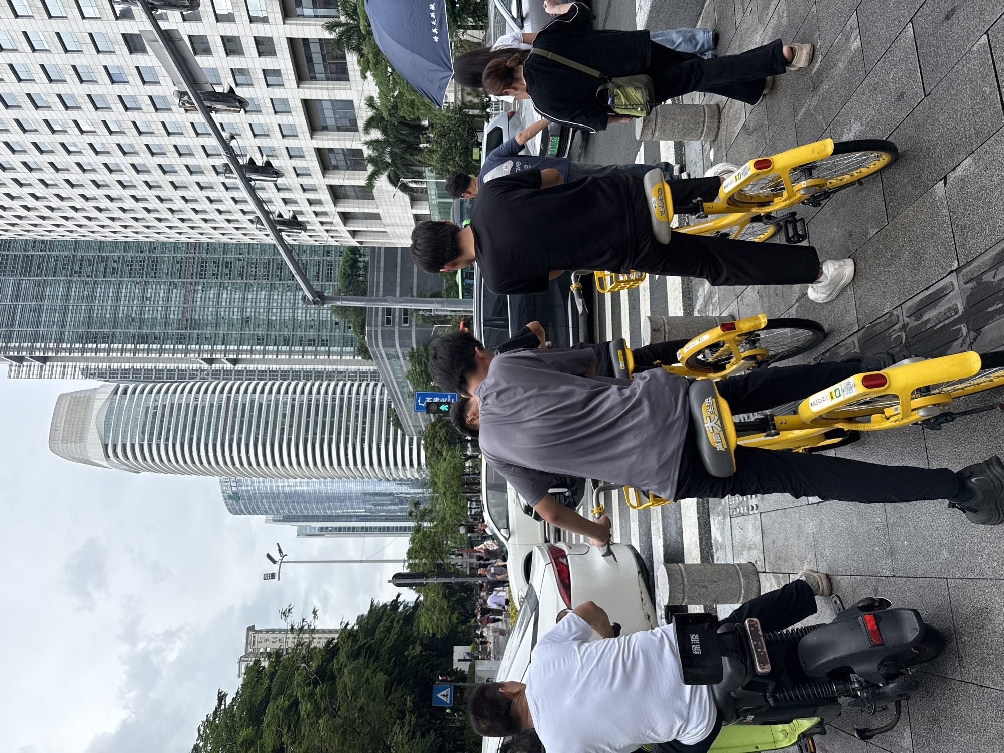 This image depicts a street scene in Tianhe District, China, characterized by modern high-rise architecture and urban infrastructure. In the foreground, four individuals are engaged with bicycles or a motorized scooter. Two yellow shared bicycles are prominent, with one ridden by a person in a grey t-shirt and black pants, and another by a person in a black t-shirt and black pants. A third yellow bicycle is partially visible to the left. A black electric scooter is being ridden by a person wearing a white t-shirt and black pants, positioned to the left of the two yellow bicycles. All individuals appear to be in motion or preparing to move, facing forward along a pedestrian crossing marked by white stripes on the grey pavement.
Behind the individuals and vehicles, the environment transitions to a busy street with vehicular traffic. White cars and a dark-colored SUV are visible on the road, suggesting a flow of traffic. In the background, the urban landscape is dominated by several imposing skyscrapers, their modern designs and reflective glass facades indicating a contemporary cityscape. Some buildings feature intricate geometric patterns, while others are sleek and cylindrical. Green trees line the left side of the street, providing a contrast to the concrete and glass structures. Overhead, traffic lights and a pedestrian crossing signal are present, along with a dark blue umbrella carried by a person further back on the right side of the scene, suggesting overcast or potentially rainy weather. The overall atmosphere is one of an active urban environment in Tianhe District, China.