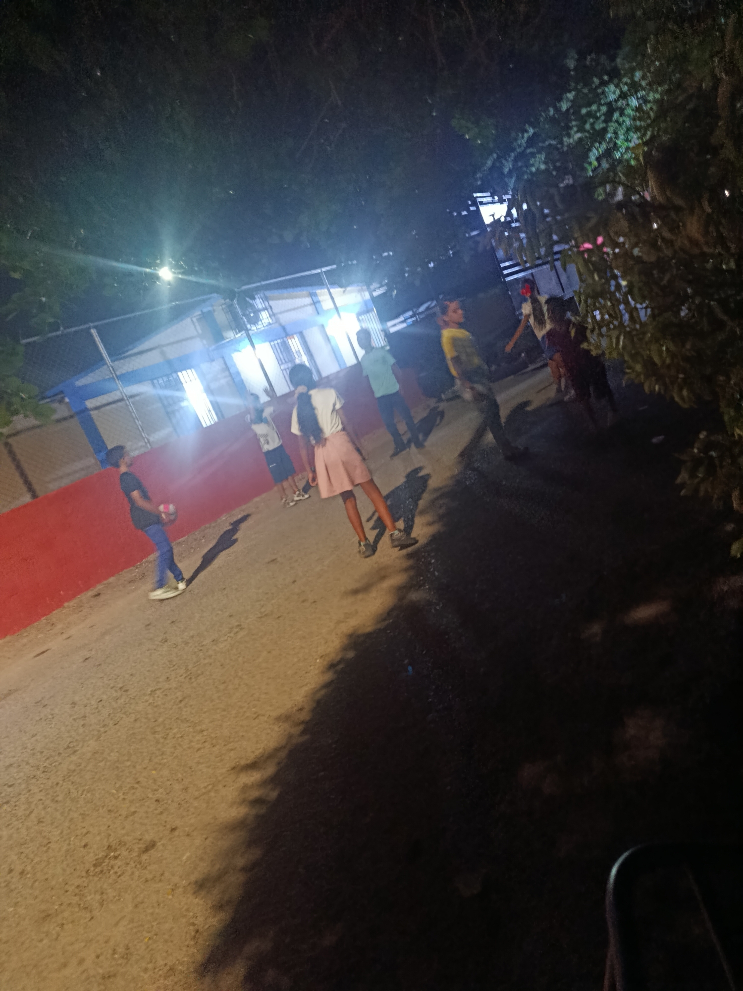 At night in Juan Griego, Venezuela, a group of children stand and play on a paved road next to a red wall and a building with a fence. A boy on the left holds a ball. Overhead lighting illuminates the scene. Trees line the edge of the road.