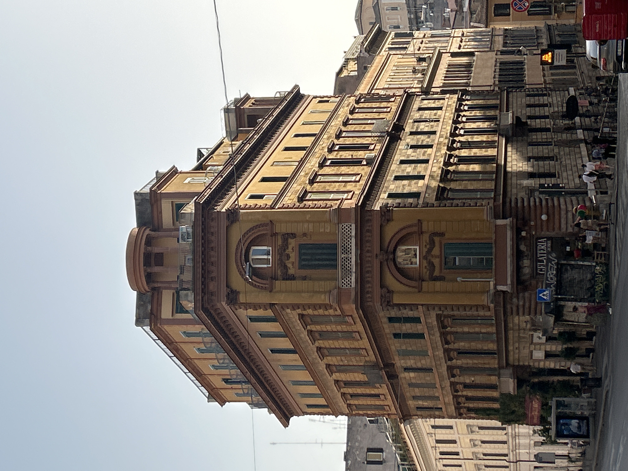 The image captures a street-level view of a tall, multi-story building in Rome, Italy, situated on a corner lot. The building features a distinct architectural style with ornate details, including balconies, arched windows, and decorative cornices. The facade is primarily painted in warm ochre and brown tones, with some sections appearing to be clad in stone.
At the ground level, there are commercial establishments, including a gelateria with a prominent sign. Several people are visible on the sidewalk and street. Some are walking, while others appear to be browsing outside the shops. A street light with a digital display is visible, along with a traffic sign indicating a pedestrian crossing. The street in the foreground is paved, and a car is parked on the right side of the image. In the background, other buildings are visible, characteristic of Rome's urban landscape, with some appearing older and in varying states of repair. The sky above is clear and pale blue, suggesting a bright, sunny day.