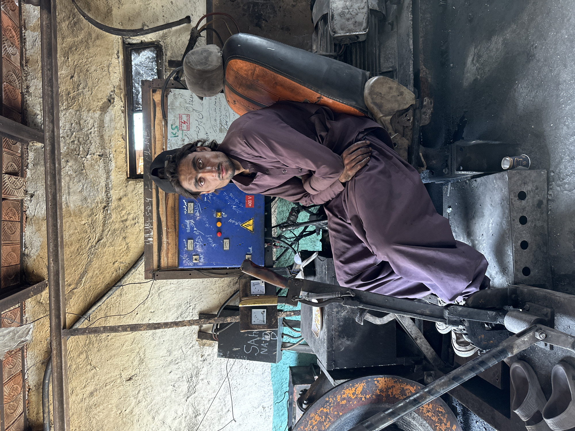 A man in a purple shalwar kameez sits on a worn, orange and black chair in a workshop in Quetta, Pakistan. He is looking directly at the camera with his arms crossed. Behind him, a blue electrical panel with meters and switches is mounted on a textured wall. Various tools and machinery are visible in the cluttered workshop, including a large metal structure to the left with an orange, rusty wheel. Several pairs of grey sandals are on the floor. The scene appears to be a workshop, possibly a metal fabrication or repair shop, given the machinery and tools present. The lighting is somewhat dim, suggesting an indoor, possibly dimly lit environment.