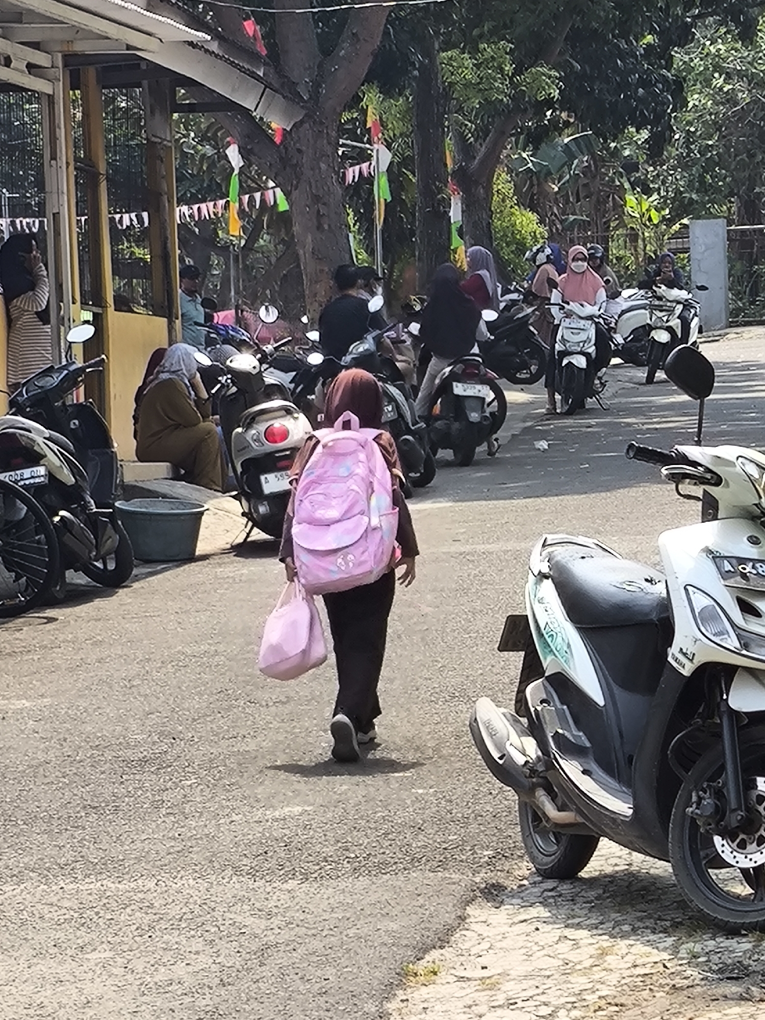The image depicts an outdoor scene on a paved road, likely in Cilegon, Indonesia, evidenced by the style of the motorcycles and the presence of small flags. In the foreground, a young girl wearing dark pants and a dark shirt with a large pink backpack walks away from the camera, carrying a pink bag in her left hand. She has a headscarf, partially obscured by her hair, and is wearing dark sneakers. Behind her, several motorcycles are parked or being ridden. To the left, a white and blue motorcycle is parked, with its front wheel and handlebars visible. Further back, a group of people, some wearing headscarves, are near more motorcycles, with a woman in a brown hijab seated on a step to the left. A large grey plastic tub is next to her. In the background, trees and foliage are visible, along with some buildings and a fence. Small, colorful flags are strung across the upper portion of the image, suggesting a festive or public event. The lighting suggests it is daytime.