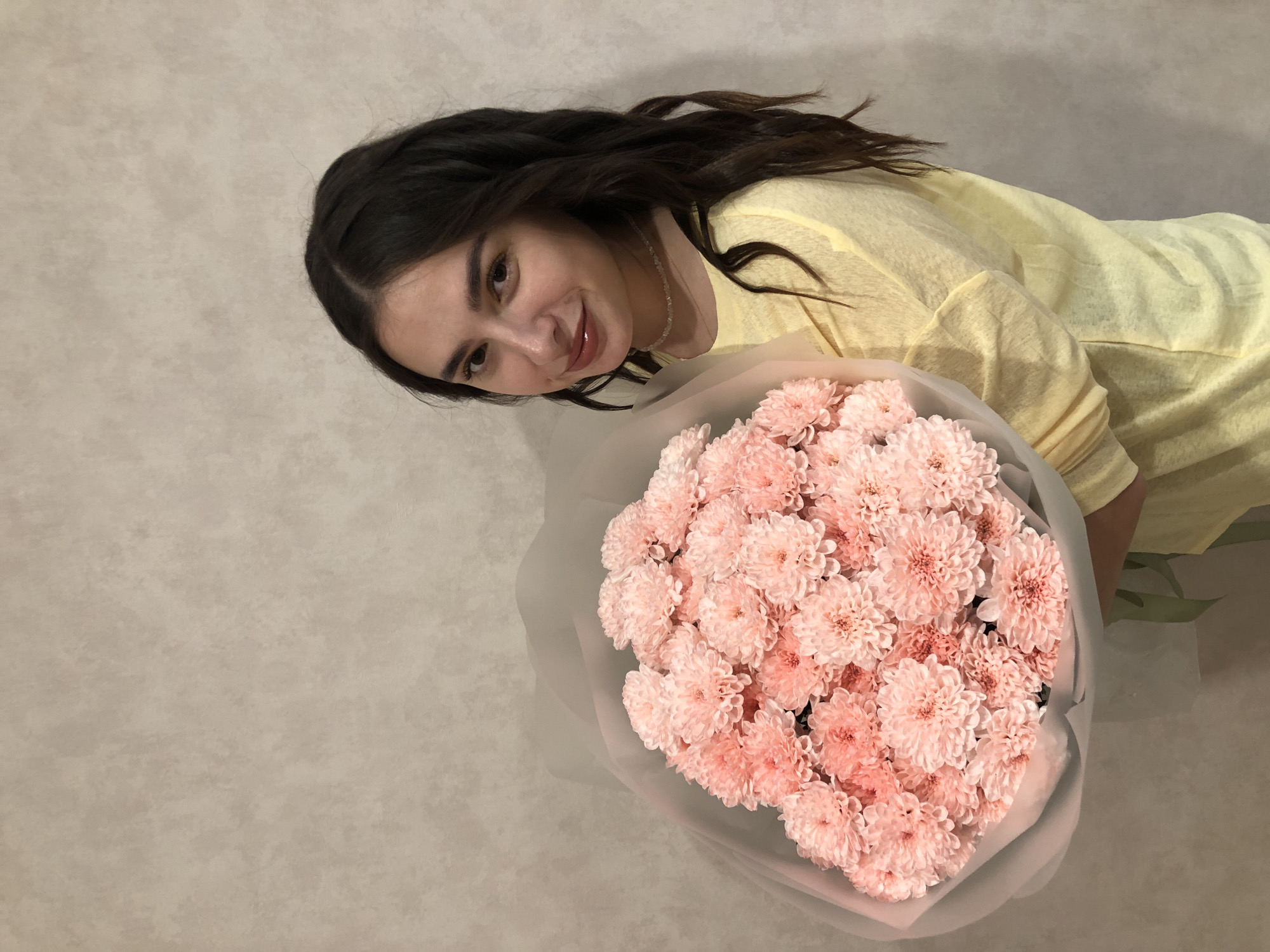 A young woman with dark hair and a yellow shirt is holding a bouquet of pink chrysanthemums. She is smiling and looking at the camera. The background is a neutral, textured wall. The lighting appears to be indoor, possibly natural light from a window. The image conveys a sense of warmth and happiness. The woman is the primary subject, with the flowers as a significant secondary element. The overall mood is cheerful and celebratory.