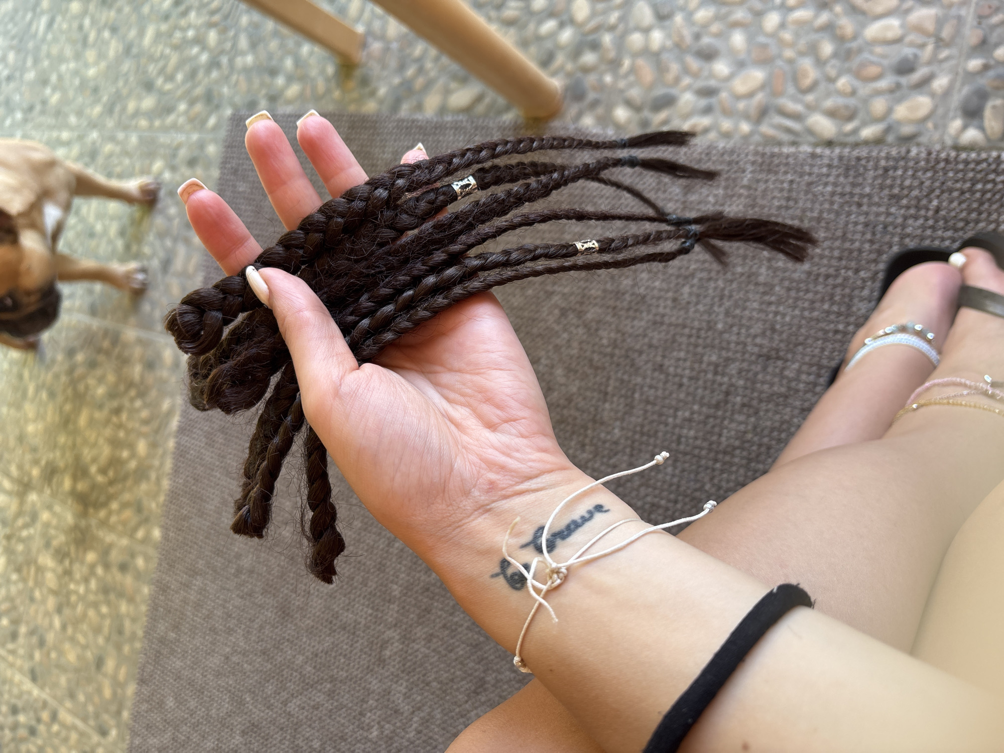 A close-up shot captures a person's hand holding a bunch of dark brown braided hair extensions. The braids are adorned with small, silver beads. The person's hand has manicured nails with a light color polish. A tattoo is visible on the person's wrist, and a thin white string bracelet is tied around it. In the background, a small, light-brown dog lies on a textured brown mat. The ground behind the dog is covered with a mosaic of small, multi-colored stones. A wooden chair leg is partially visible in the upper portion of the frame. The lighting suggests it is daytime, possibly outdoors given the stone flooring and mat. The scene appears relaxed and casual.