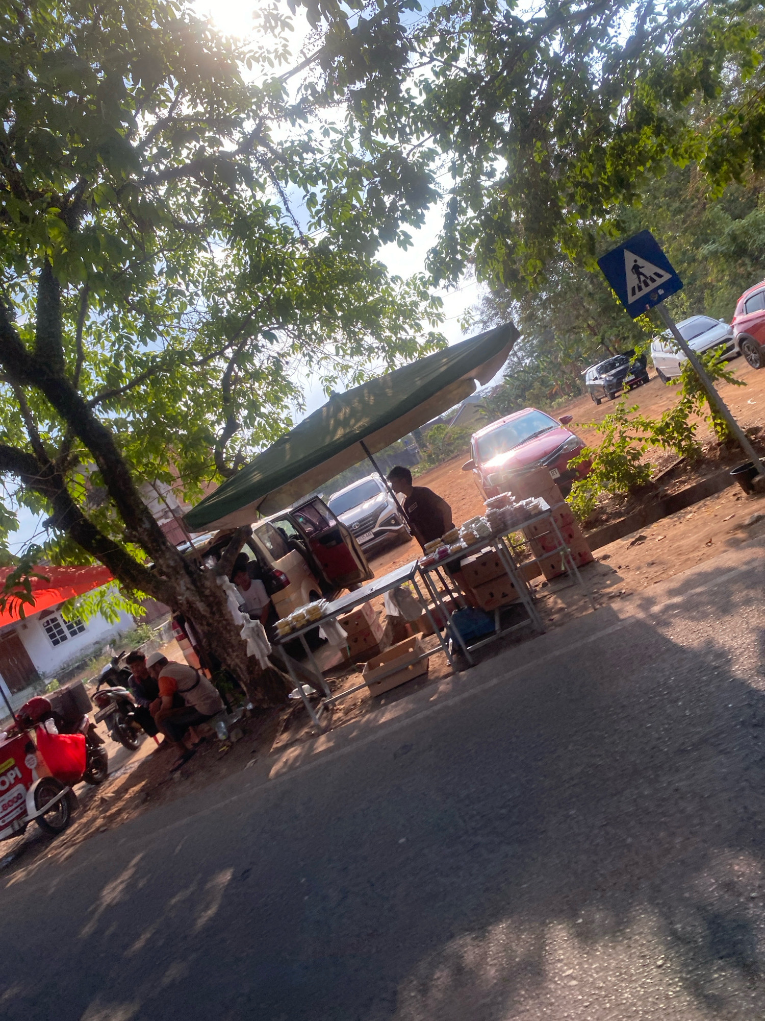 The image depicts a street scene in Metro, Indonesia, characterized by a roadside stall and several vehicles. In the foreground, a paved road is visible with dappled sunlight and shadow patterns cast by overhead foliage. To the left, a red and white food cart with visible text indicating "OPI" and "300" is parked, accompanied by a motorcycle. Further back, a makeshift stall with a green canopy displays various food items and boxes.
Several people are present at the stall. A man in a dark shirt is interacting with the stall's offerings. Two other men, one wearing a white cap, are seated on a motorcycle near a tree. A beige van with open doors and a red car are parked behind the stall, and other vehicles, including a white car and a black SUV, are visible further in the background. A pedestrian crossing sign is also present on the right side of the image. The environment appears to be a semi-urban or suburban area with trees and vegetation bordering the road. The sun is shining brightly, suggesting it is daytime.