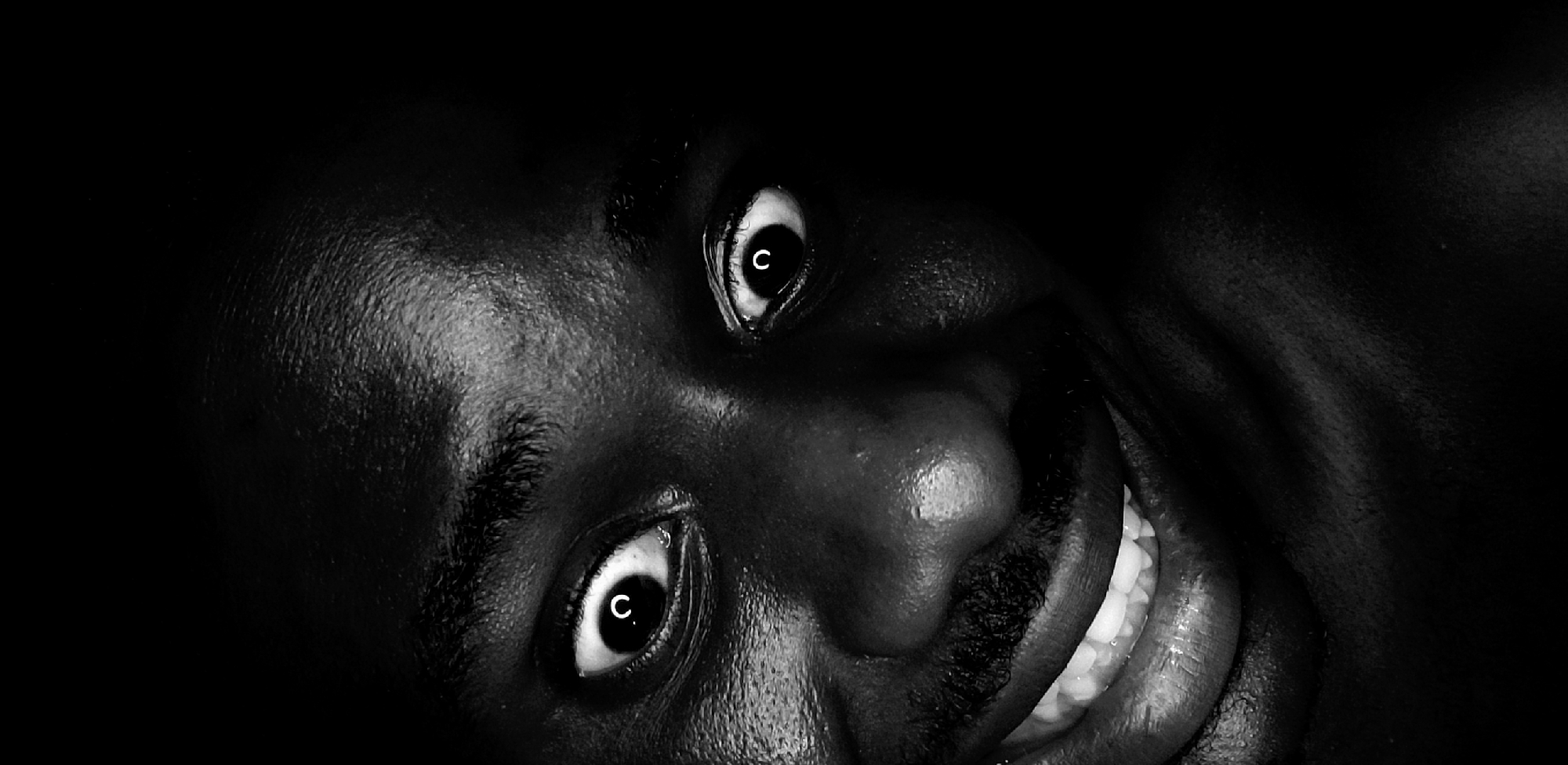 An extreme close-up, high-contrast, black and white portrait captures the intensely focused face of a person, likely a man given the facial hair, set against a completely dark background. The image, originating from Narayi, Nigeria, features dramatic lighting that highlights the subject's features.
The central focus is on the face, which is angled slightly and occupies most of the frame. The subject's eyes are wide open, their whites prominent, and they stare directly at the viewer with an unsettling intensity. A notable detail is the distinct white letter 'C' visible within the dark pupil of each eye, appearing as a reflection or overlaid element.
Below the eyes, a mustache is visible, and the person's mouth is open in a wide smile or grin, revealing a full set of bright white teeth. The dark skin appears slightly reflective, catching the light and emphasizing its texture. The background is uniformly black, offering no environmental cues. The overall expression conveys a mix of intensity, directness, and perhaps a playful or mischievous amusement.