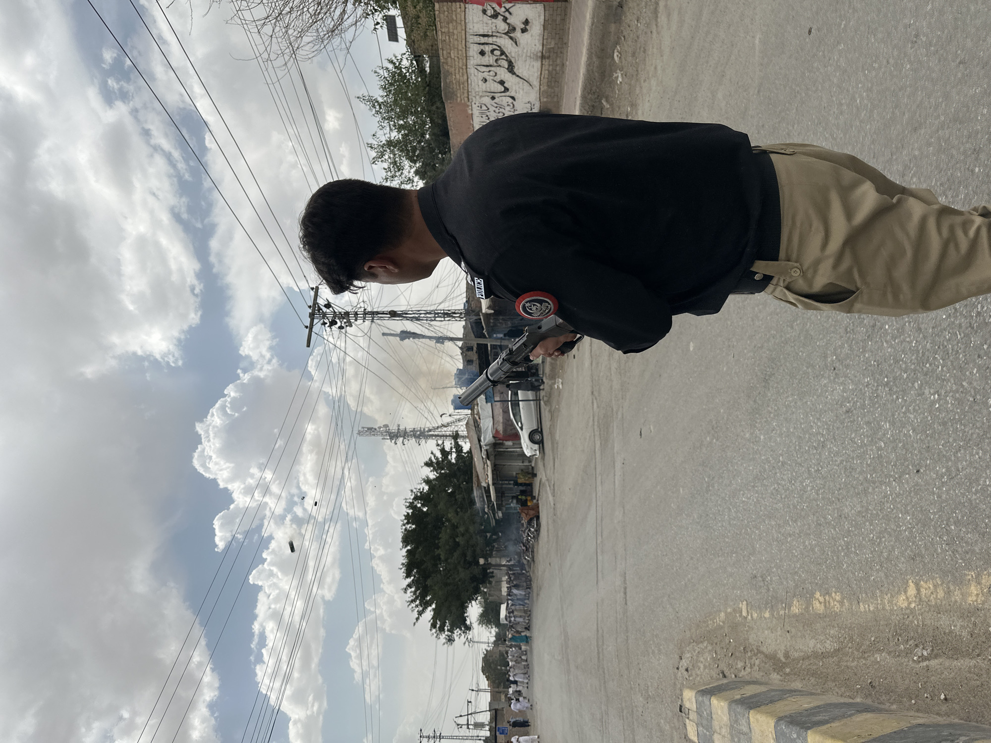 In an outdoor, urban environment under a cloudy sky, a police officer wearing a black uniform and tan pants stands facing away from the camera, holding a firearm. The officer's uniform has patches on the sleeve and the word "POLICE" visible on the collar. The background depicts a street lined with buildings and shops, a white car parked, and a group of people gathered. Smoke is rising from an area further down the street. Electrical wires and utility poles are prominent features of the scene. The road surface is asphalt, with a yellow and black striped speed bump visible in the foreground. Graffiti is present on a wall in the background.
