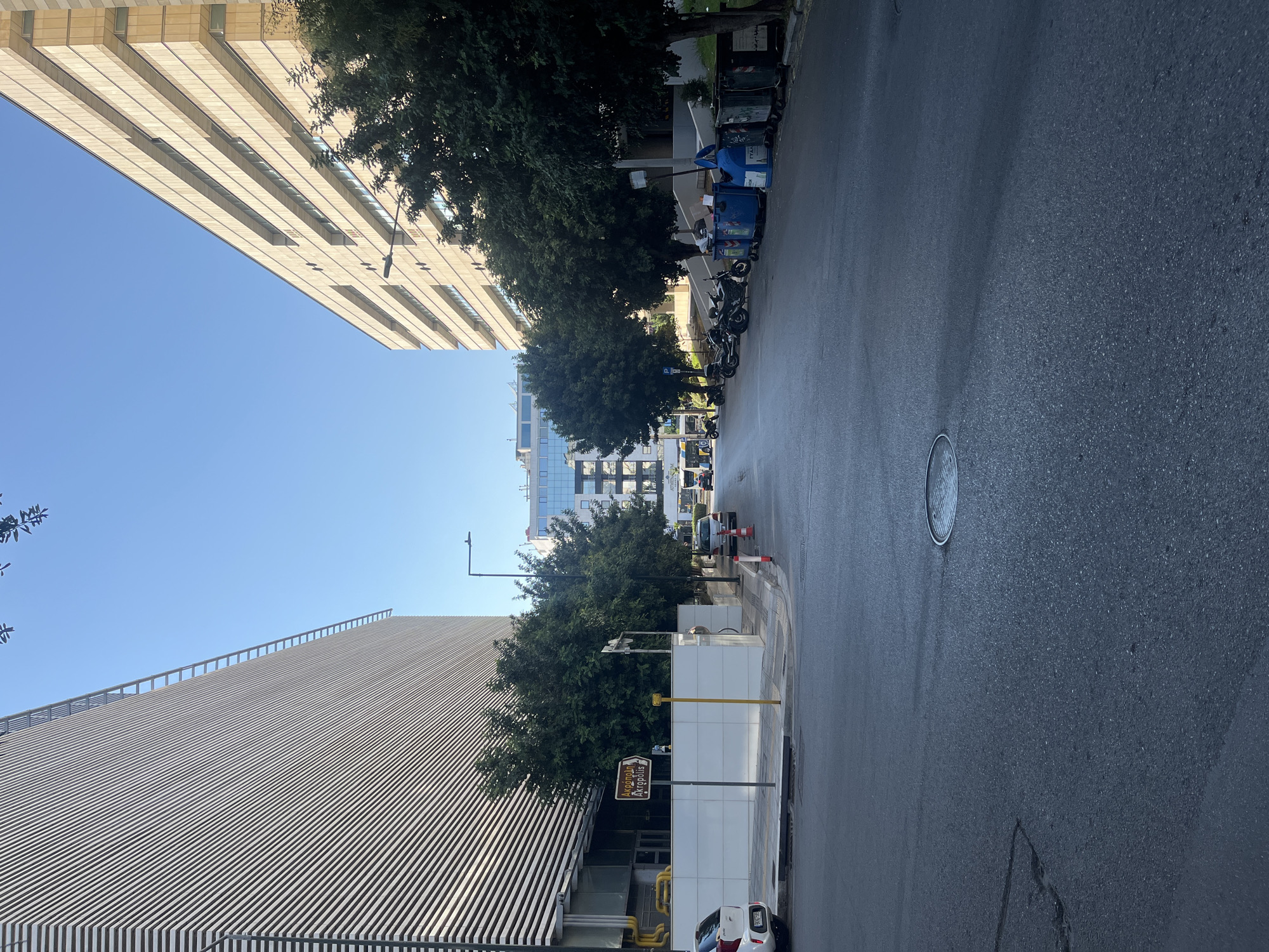 The image depicts a street scene in Athens, Greece, likely in a commercial or business district. On the left, a large, modern building with a ribbed facade dominates the frame. To the right, another multi-story building with a beige, layered exterior is visible. The street itself is paved and shows signs of wear, with a manhole cover prominently in the foreground.
Several parked motorcycles and scooters are lined up along the right side of the street. Adjacent to them are several large blue and gray trash bins. Trees with full green foliage are planted along the sidewalk, providing shade.
In the middle distance, the street extends towards more buildings, one of which appears to have a glass facade. Traffic cones are visible on the side of the road, and a few cars are present. A street sign in Greek, pointing to "Akropolis," is mounted on a pole on the left side of the street. The sky is clear and blue. The overall impression is one of a bright, sunny day in an urban environment.