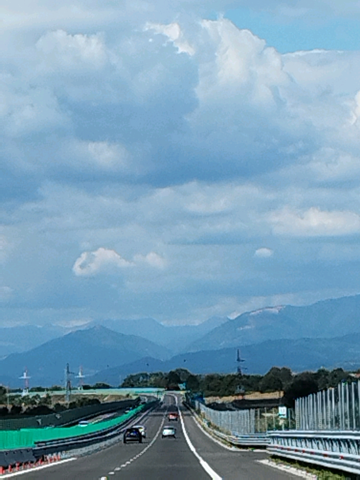 The image depicts a highway with several cars driving on it. The highway is flanked by sound barriers and guardrails. In the background, there are mountains and trees, and the sky is filled with clouds. The scene appears to be a typical highway landscape on a cloudy day. There are no visible people in the image. The location in the city of Anghinești, Romania, is not indicated or discernible from the visual content of the image.