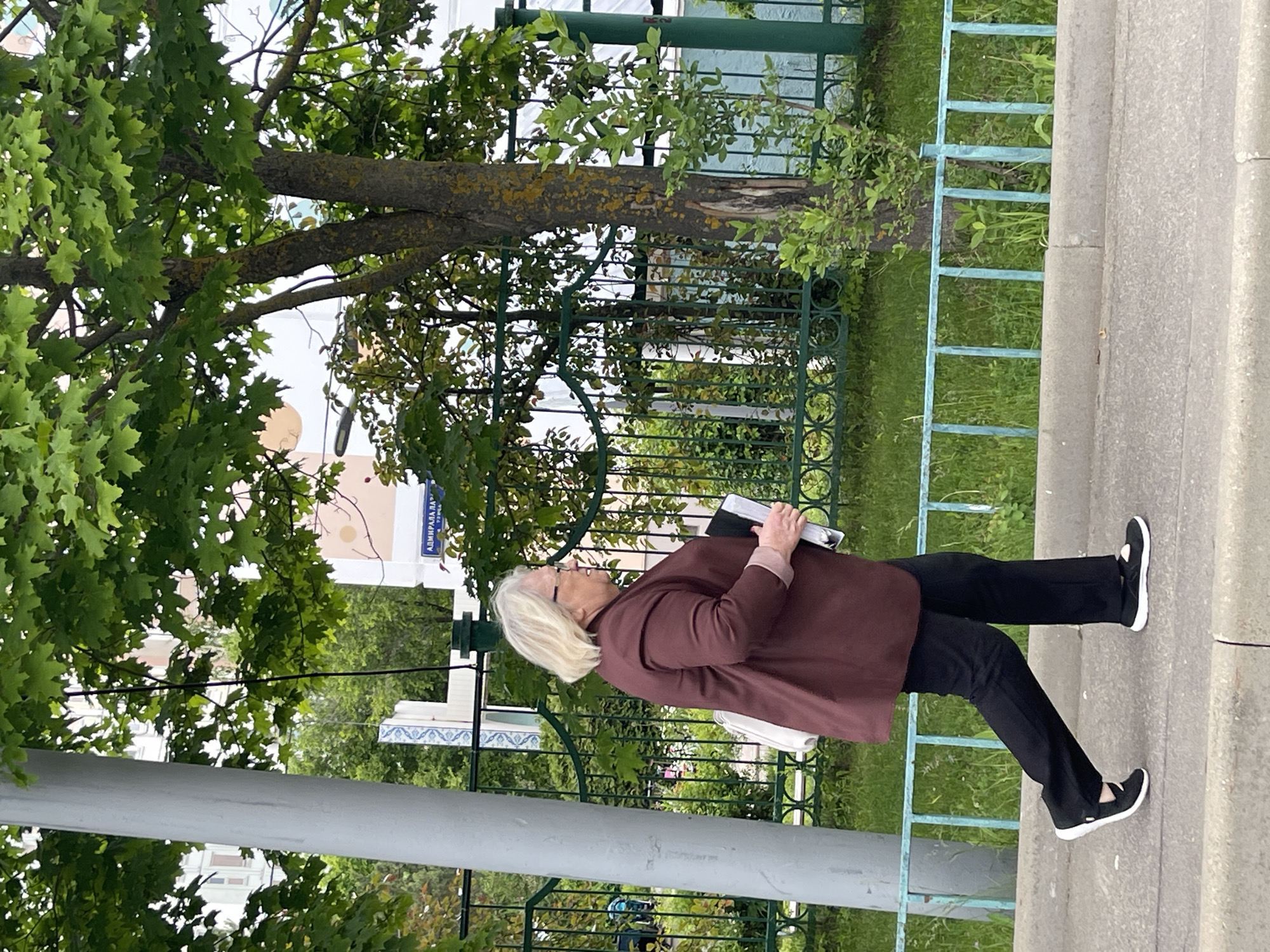 An eye-level medium shot presents an elderly woman walking on a sidewalk in Moscow, Russia. She is dressed in a maroon jacket, black pants, and black shoes with white soles. She holds a book and a white bag. The background features lush greenery, a green metal fence, and street signs reading "Admiralala". The scene is outdoors on a day with cloudy overcast weather.