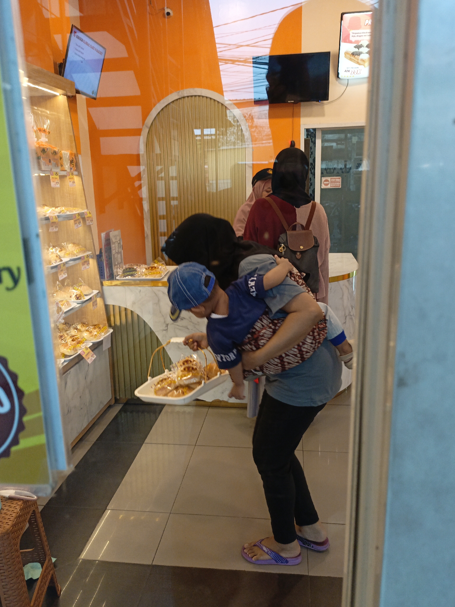 In East Jakarta, Indonesia, a scene unfolds within a bakery. A woman, her face obscured by a dark headscarf, is carrying a young child, who is facing away from the camera and wearing a blue cap. The child is being held in a wrap tied around the woman's torso, and is reaching for a white tray filled with pastries. The woman herself is wearing black pants and purple flip-flops. Behind her, two other women, also wearing headscarves, are visible, one in a pink hijab and the other in a maroon sweater with a brown backpack. The background features shelves stocked with packaged bread, an orange wall, and a curved entryway with wooden louvers. A small television is mounted on the orange wall. The floor is tiled in a checkerboard pattern of light and dark grey squares.