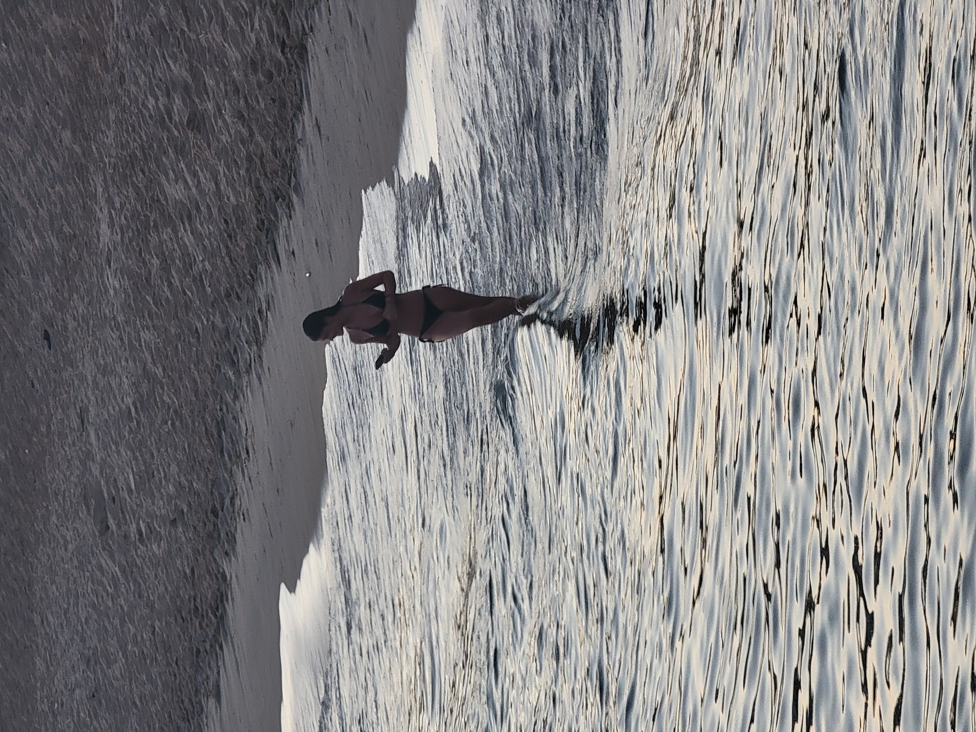 A woman in a black bikini is walking in shallow water near a sandy beach. The woman is facing left and looking at her phone. The water is rippling and reflecting the light. The sand is wet from the waves. The scene appears to be a beach in Quarteira, Portugal, possibly during dusk or dawn due to the lighting.