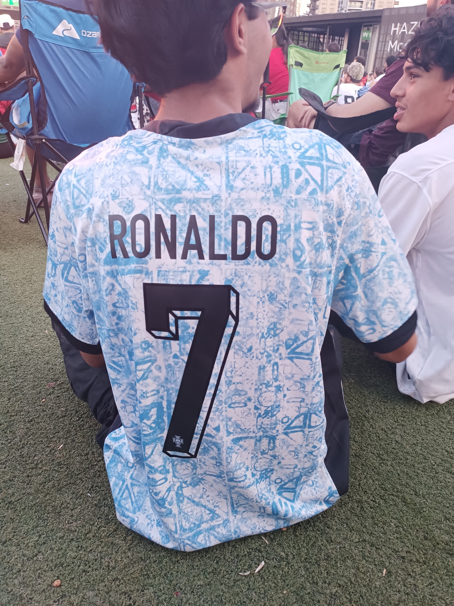 The image shows a person from behind wearing a light blue and white patterned soccer jersey with "RONALDO" and the number "7" printed on the back in black. The person is sitting on a patch of artificial turf. To the left of the person, a blue folding chair with "OZARK" printed on it is visible. In the background, other people are seated, and some buildings and a banner with the word "HAZ" are visible. The overall setting appears to be an outdoor event. This scene is not identifiable as being specifically in Mississauga, Canada.
