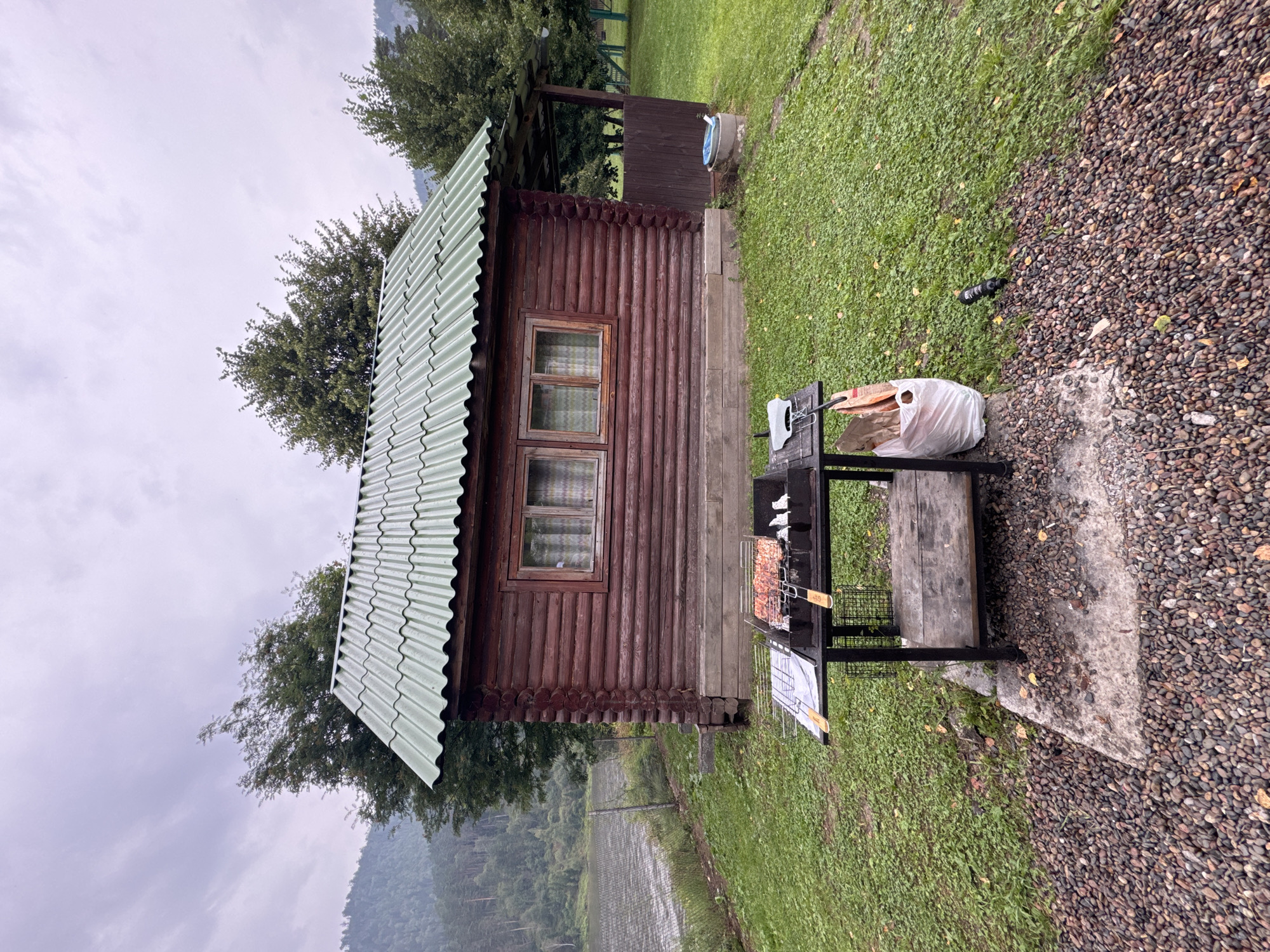 The image depicts an outdoor scene in Усть-Мана, Russia, featuring a rustic log cabin with a green corrugated roof. In the foreground, a black metal barbecue grill, loaded with raw meat on skewers, sits on a concrete pad surrounded by gravel. Beside the grill, a white plastic bag and a black tool are visible on the grass. The cabin is situated on a grassy lawn, with trees and a body of water visible in the background. The sky is overcast, suggesting a cloudy day.
