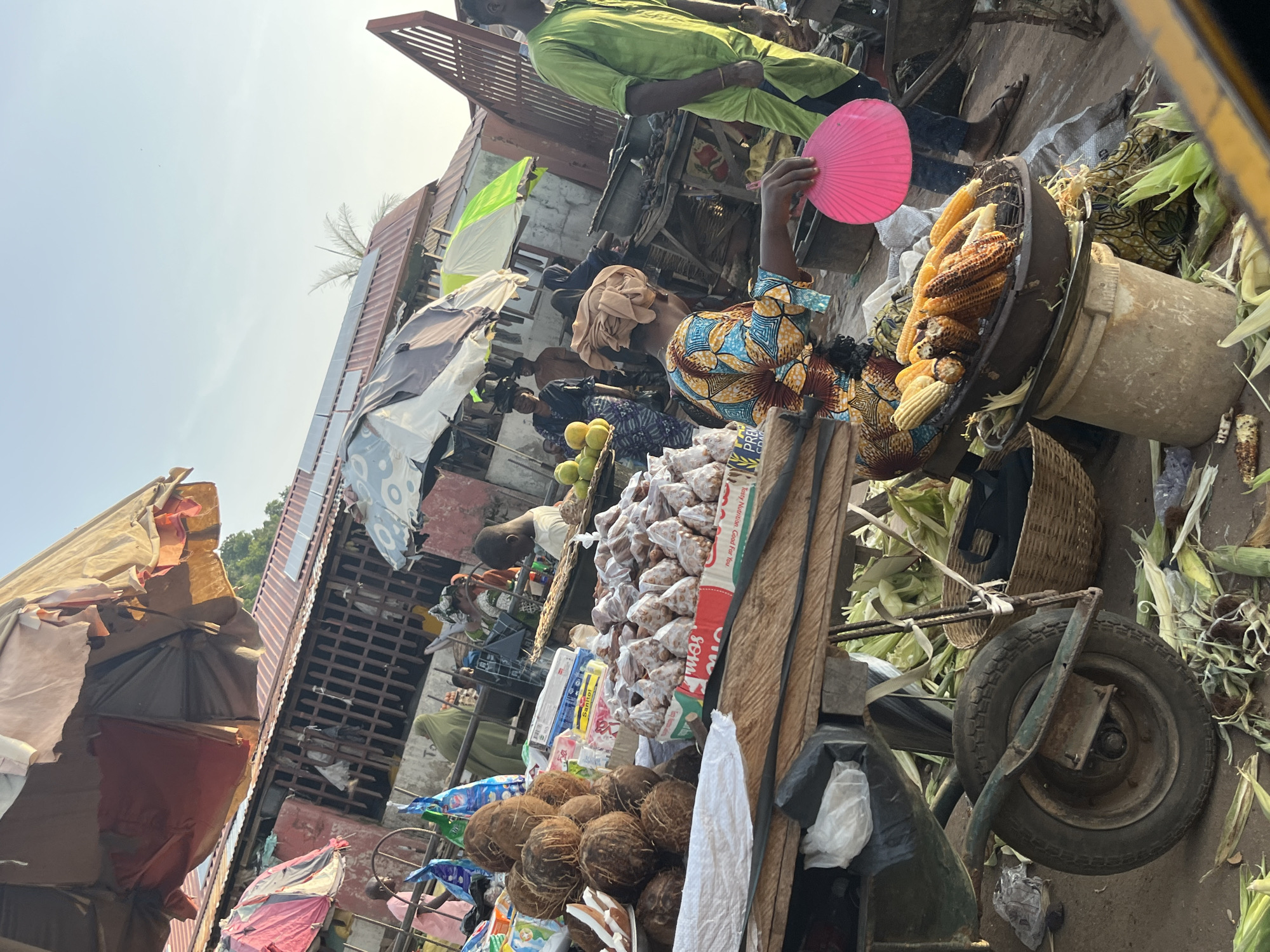 This image captures a bustling outdoor market scene in an urban environment, likely in a tropical or subtropical climate, given the presence of coconuts and corn. The photograph is taken from a slightly elevated perspective, looking down at a busy marketplace.
In the foreground, a wooden cart is laden with goods. Prominently displayed are several whole coconuts, some of which appear to have been split open, revealing their white flesh. Next to the coconuts, there are numerous small, individually wrapped food items, possibly snacks or sweets, packaged in clear plastic bags and arranged on a tray. Below this, a large basket of roasted or grilled corn on the cob is visible, resting on a cylindrical object, possibly a bucket. More corn husks and cobs are scattered on the ground around the cart.
A woman, wearing a brightly colored, patterned dress and a headscarf, is the central figure in this foreground area. She is holding a bright pink fan and appears to be attending to her stall. Her gaze is directed downwards and slightly to her right.
Behind the cart, the market unfolds with more stalls and people. Umbrellas, some tattered and weathered, provide shade over various displays of goods. Several people are visible, some in the process of selling or buying, and others simply present in the market environment. Clothing styles suggest a diverse group of individuals. The structures in the background appear to be simple, possibly temporary market stalls or small buildings with corrugated metal roofs, some of which are fitted with solar panels, indicating a blend of traditional and modern elements. The overall atmosphere is one of vibrant commerce and daily life.