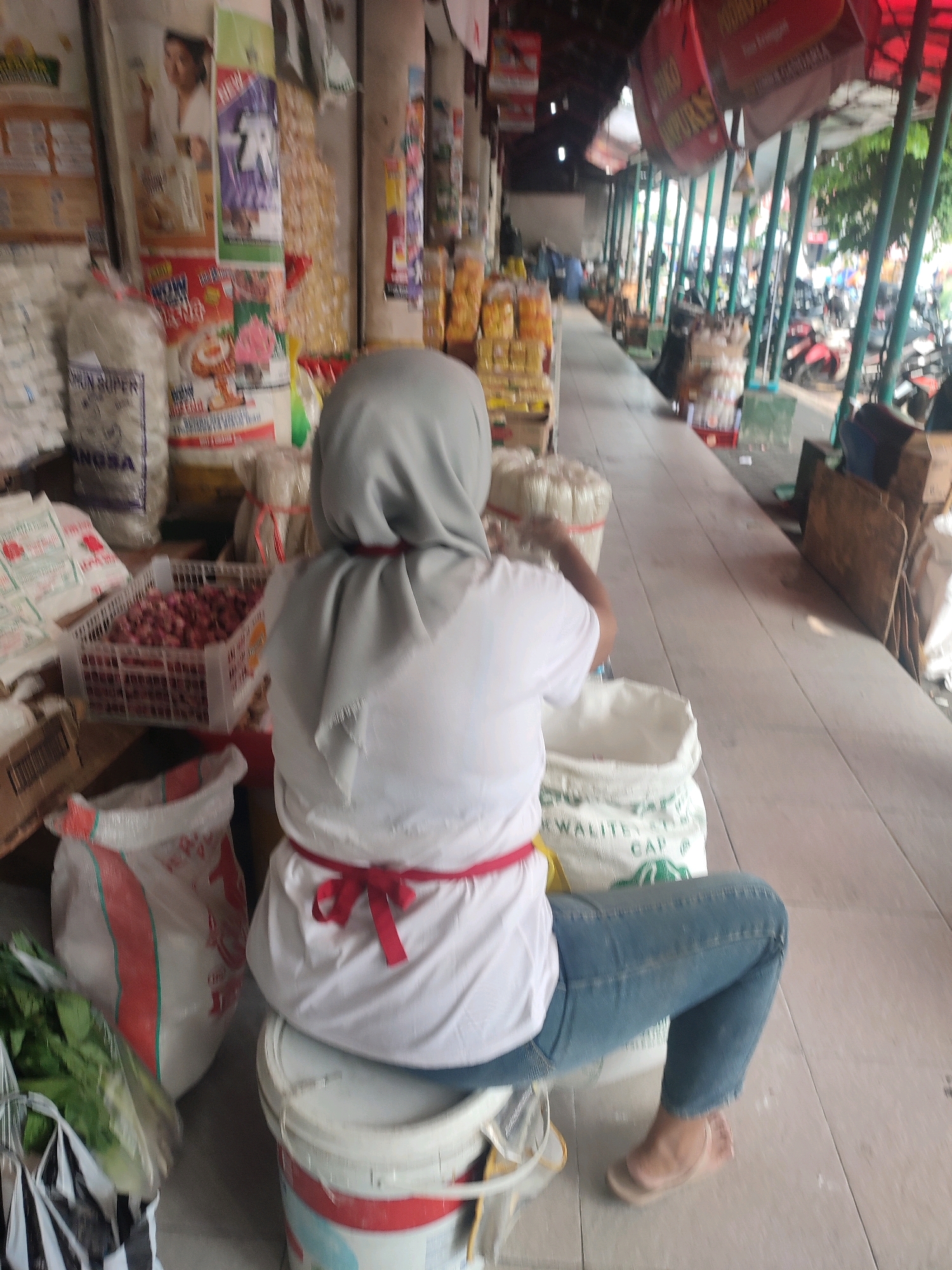 A medium shot captures a person from behind, wearing a light grey hijab, a white t-shirt tied with a red sash, and blue jeans. They are seated on a white bucket, positioned between two large sacks, one white with a red stripe and the other predominantly white with green lettering. The person's right hand is reaching into the white sack in front of them. The setting appears to be an outdoor market, with a tiled walkway and stalls lining the right side of the frame. These stalls are adorned with various products, including bags of grains, snacks, and produce. In the background, several motorcycles are visible, suggesting a busy marketplace. The lighting indicates daytime. This scene is consistent with the environment of Beringharjo Market, a well-known traditional market in the city of Yogyakarta, Indonesia.