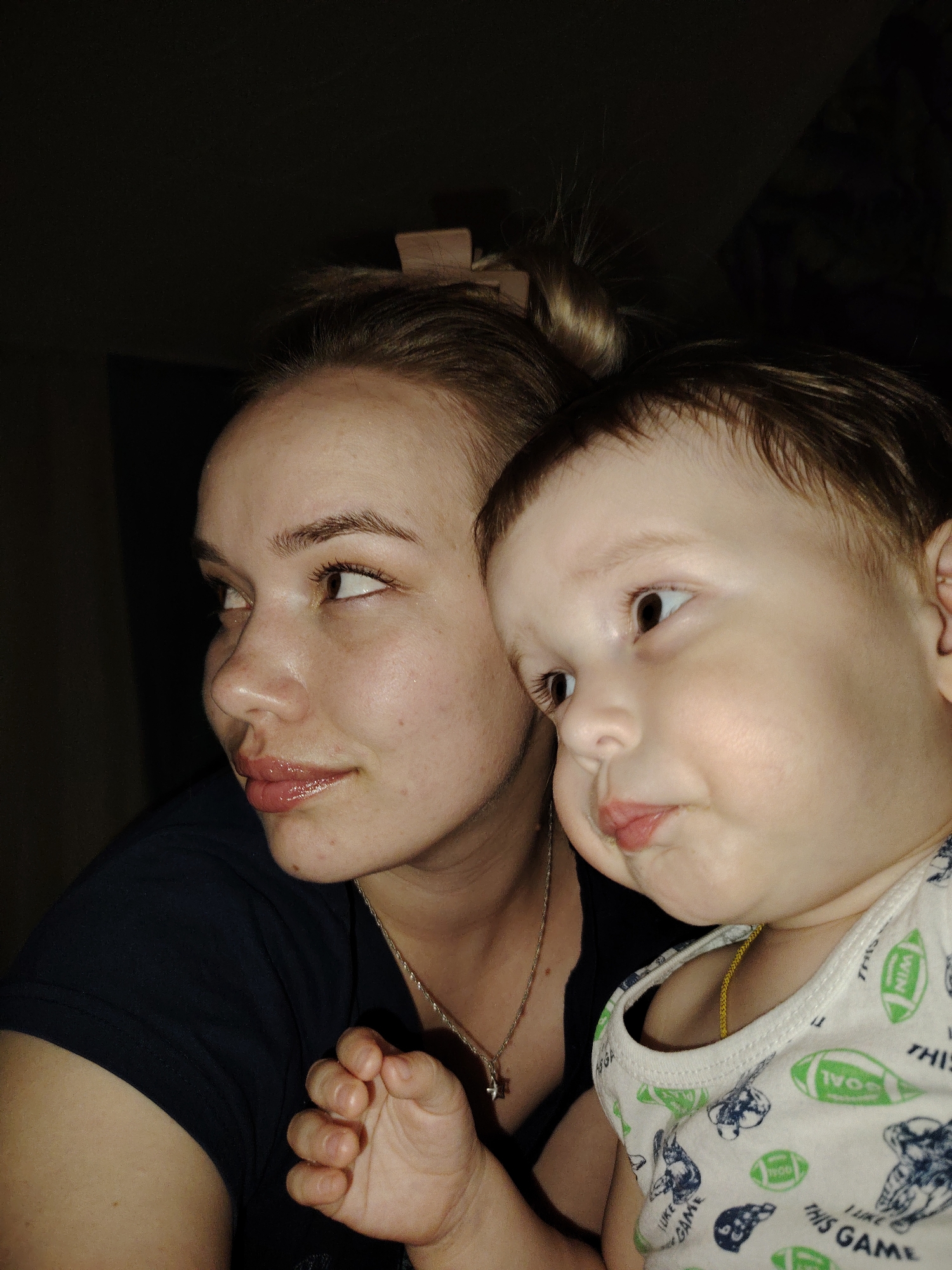 An indoor scene features two individuals positioned in close proximity, illuminated by an off-camera light source in a predominantly dark environment. No identifying markers for the city of Марковский, Russia, are discernible.
On the left, an adult female with light skin, brown hair styled in a messy bun secured with a light-colored clip, and brown eyes, is depicted in profile, looking towards the left side of the frame. Her lips are slightly parted and appear glossy. She is wearing a dark, possibly navy or black, short-sleeved top and a thin silver necklace with a small cross pendant. Minor skin blemishes are visible on her chin and forehead.
To her right, partially obscured by the female's head and shoulder, a young child, likely a toddler, also with light skin, dark brown hair, and brown eyes, is positioned. The child's face is in three-quarter profile, looking in the same direction as the female, with lips slightly pursed. The child is wearing a white sleeveless top patterned with green footballs, the words "THIS GAME," and what appear to be cartoon animal figures (bunnies). A thin gold chain is visible around the child's neck. The child's left hand, clenched into a fist, rests near the adult female's upper arm.
The background is uniformly dark, indicating low light conditions and providing no additional contextual details.