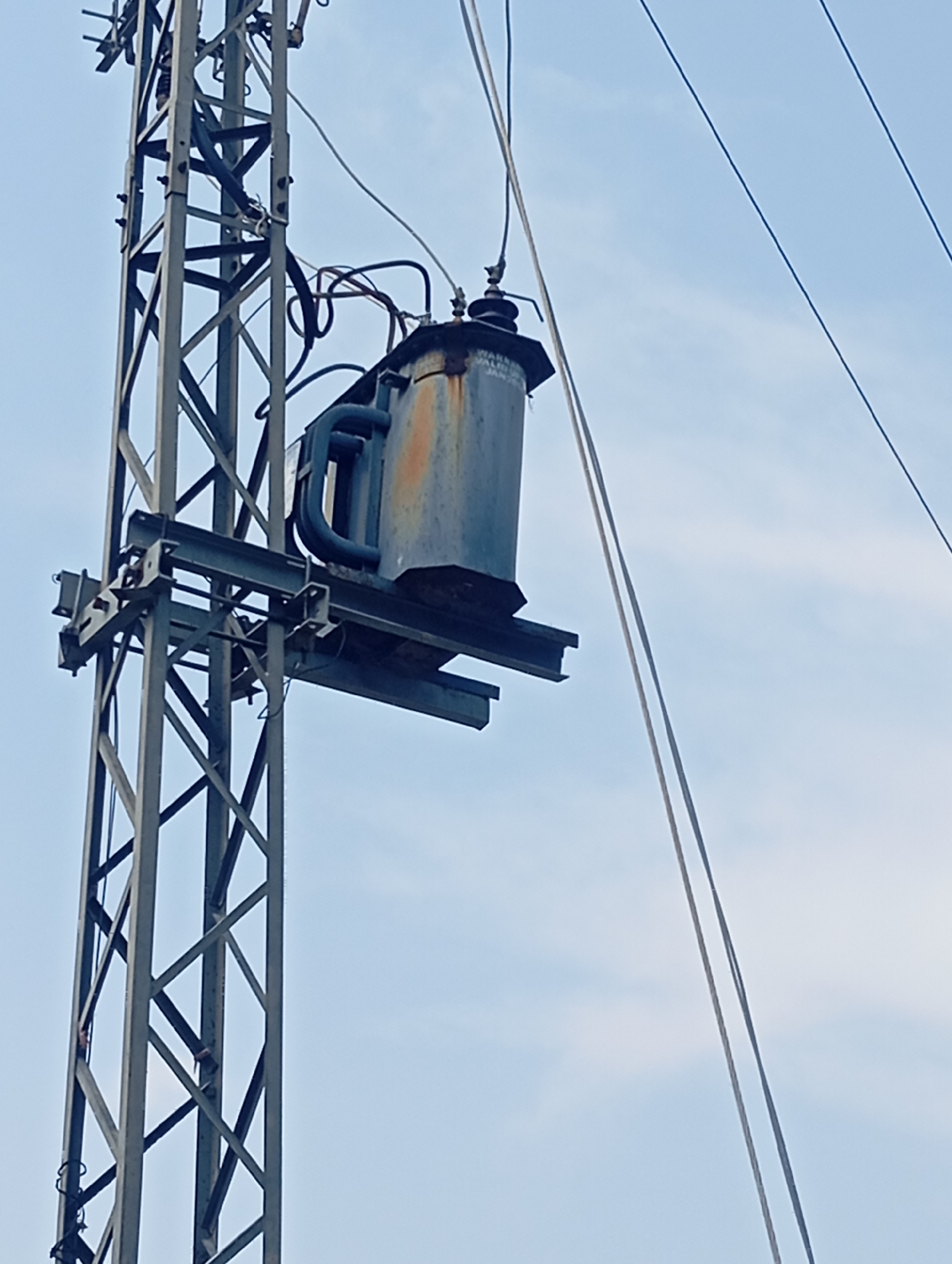 An outdoor, medium shot captures a power transformer mounted on a metal utility pole against a pale blue, cloudy sky. The transformer, a weathered, gray cylindrical object with some rust on its upper portion, is positioned to the right of the frame. It is connected by thick black cables to the utility pole, which is a lattice structure of gray metal bars. Two power lines extend diagonally from the upper right towards the left of the frame, also connected to the pole. The overall scene suggests a functional, albeit aged, electrical infrastructure in an outdoor setting. The image does not contain any people or signs of human activity beyond the installation of the electrical equipment.