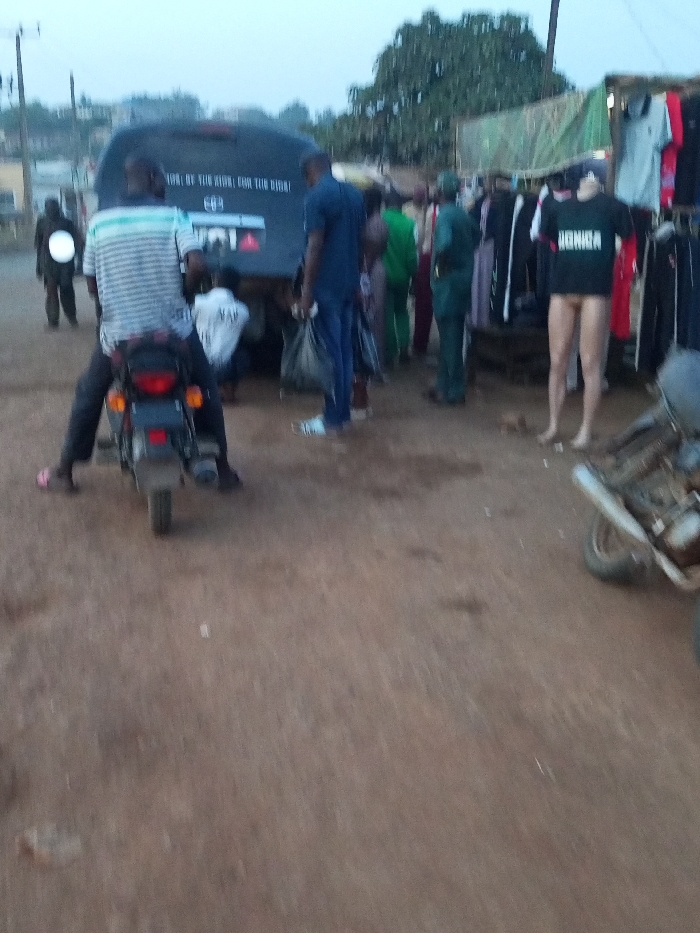 A bustling outdoor market scene unfolds. A motorcyclist waits near a black van with text on its rear. Clothes are displayed on a makeshift stand under a green tarp. Mannequins, one wearing a black shirt and the other without clothes, stand beside the rack. People mill around. Another motorcyclist approaches with their headlight shining. The ground is dirt and the sky is overcast.