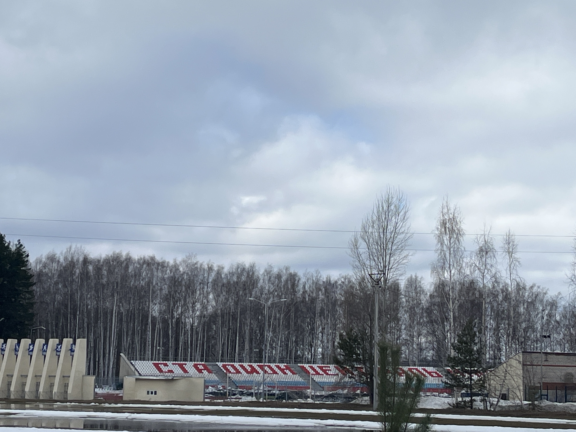 The image shows a stadium in Chaikovsky, Russia, situated behind a line of bare trees. The seating area is visible, with text in Russian written across the seats in red and white. A building is adjacent to the stadium. In the foreground, there is evidence of melting snow. The sky is overcast. Power lines are visible above the trees. The architectural style of the stadium and surrounding structures appears utilitarian and typical of post-Soviet design.
