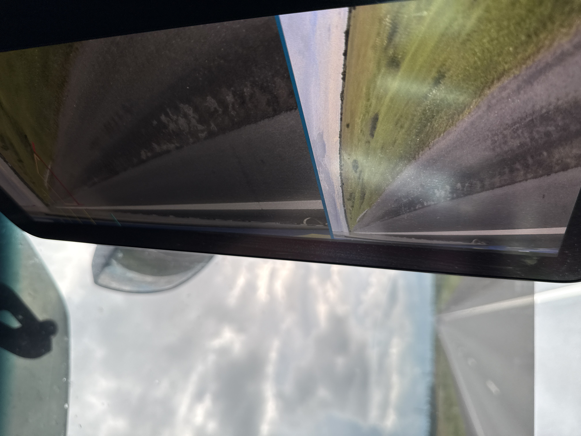 The image shows a view from inside a vehicle, looking out through the windshield and into a side mirror which displays a feed from a backup camera. The windshield reflects a cloudy sky and a blurred view of a road stretching into the distance. The side mirror shows a split image: the upper portion displays a section of the road ahead with painted lane lines, while the lower portion shows a wider, panoramic view of a grassy field beside a paved road, with faint red and yellow guide lines indicating the camera's view. The scene is consistent with a highway environment in an open landscape. There is no indication of the city of Kipuya, Russia.