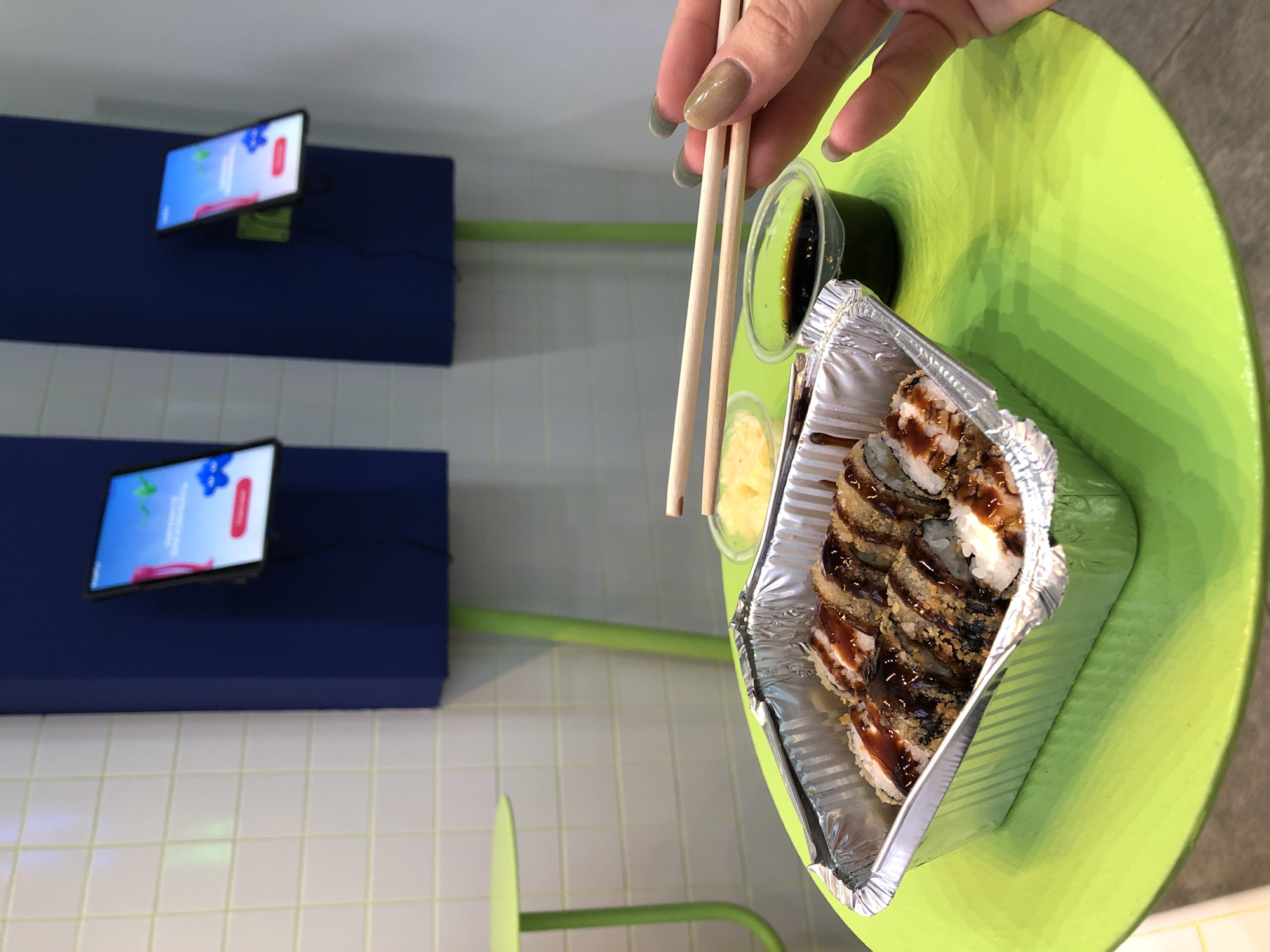 A hand with light brown-green nail polish is holding chopsticks over a foil container of sushi rolls on a bright green circular table. The sushi appears to be deep-fried and drizzled with a dark sauce. To the right of the sushi is a small clear container with dark liquid, likely soy sauce, and to the left is a small container of yellow pickled ginger. In the background, two blue vertical panels are mounted on a white tiled wall, each displaying a tablet screen with a colorful interface. Green metal table legs are visible supporting small green circular tables. The scene appears to be inside a modern cafe or restaurant, possibly located in Chaikovsky, Russia.