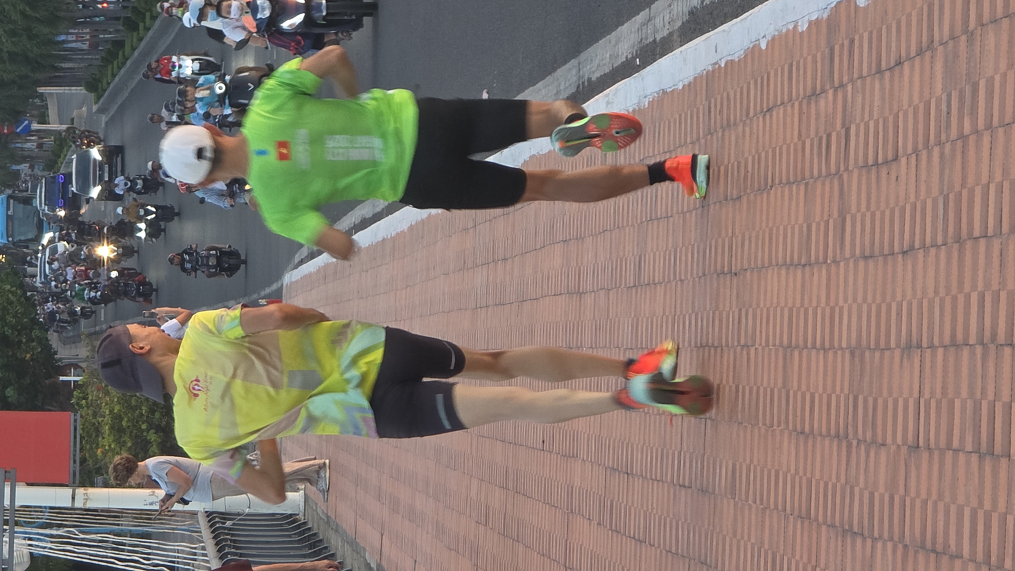 The image depicts two male runners jogging on a reddish-brown interlocking brick sidewalk, identified as part of a bridge or elevated structure in Nha Trang, Vietnam. The lead runner, positioned closer to the viewer and slightly to the left, wears a dark baseball cap, a yellow and multi-colored tie-dye t-shirt, black running shorts with a light reflective stripe on the left leg, and multi-colored running shoes (predominantly orange, green, and yellow). His face is partially visible in profile, showing a slight smile. The second runner, behind and to the right of the first, wears a white baseball cap, a bright green t-shirt featuring a Vietnamese flag patch and a white number '1' on the back, black running shorts, black socks, and orange and green running shoes.
To the immediate left of the runners, a light-colored metal railing borders the sidewalk. A woman with short, light brown hair stands near this railing, observing her mobile phone. Her attire consists of a light grey t-shirt and light-colored pants.
To the right of the runners, a multi-lane asphalt road is separated from the sidewalk by a white line. The road is densely populated with a mix of traffic, primarily numerous motorcycles and scooters, along with several cars and a blue bus visible in the distance. All vehicles are traveling in the same direction, away from the viewer, indicating a busy urban thoroughfare. Many motorcycles have illuminated headlights. The ambient lighting suggests either early morning or late afternoon/dusk. Trees and foliage line the far side of the road.