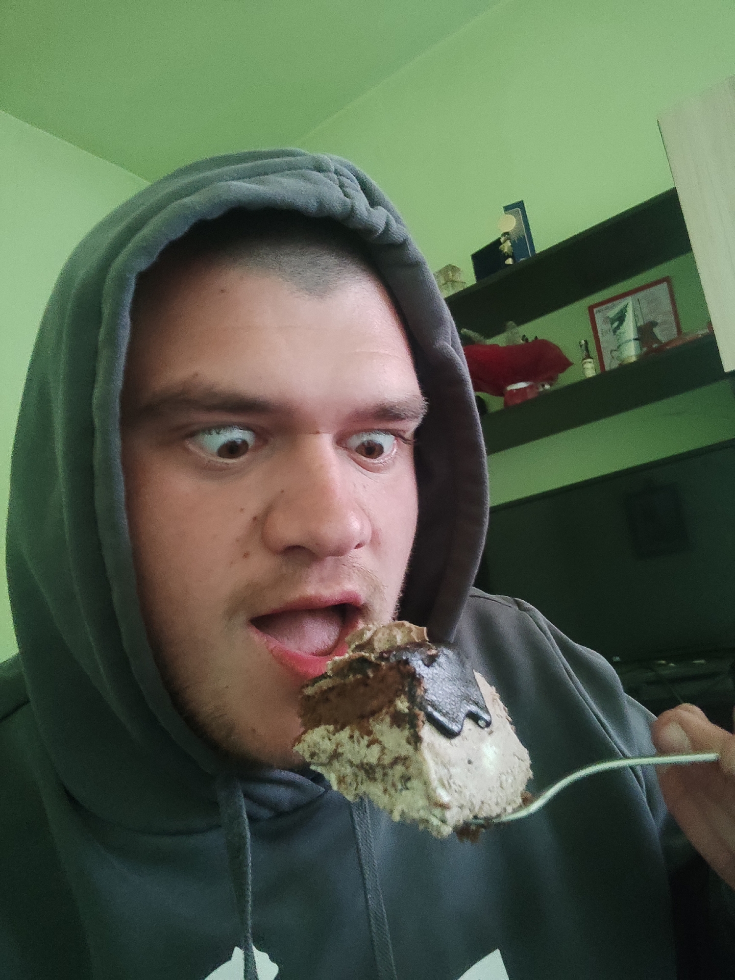 A man with short-cropped hair wearing a gray hooded sweatshirt holds a fork with a piece of cake with chocolate sauce in front of his mouth. He has his eyes wide and mouth open, seemingly about to eat the cake. The background includes a green wall and a dark shelf with various objects. It is not possible to identify the location as Burgas, Bulgaria from the image.