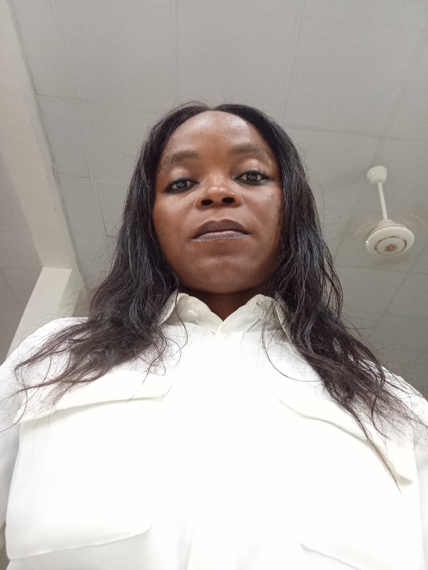 A low-angle, medium shot shows a Black woman with long, dark hair, wearing a white collared shirt. She is looking directly at the camera with dark eye makeup. Her lips are slightly parted and appear to have a metallic sheen. The background is a lightly textured ceiling with fluorescent lighting and a white ceiling fan visible in the upper right corner. The image was taken in Kason, Nigeria.