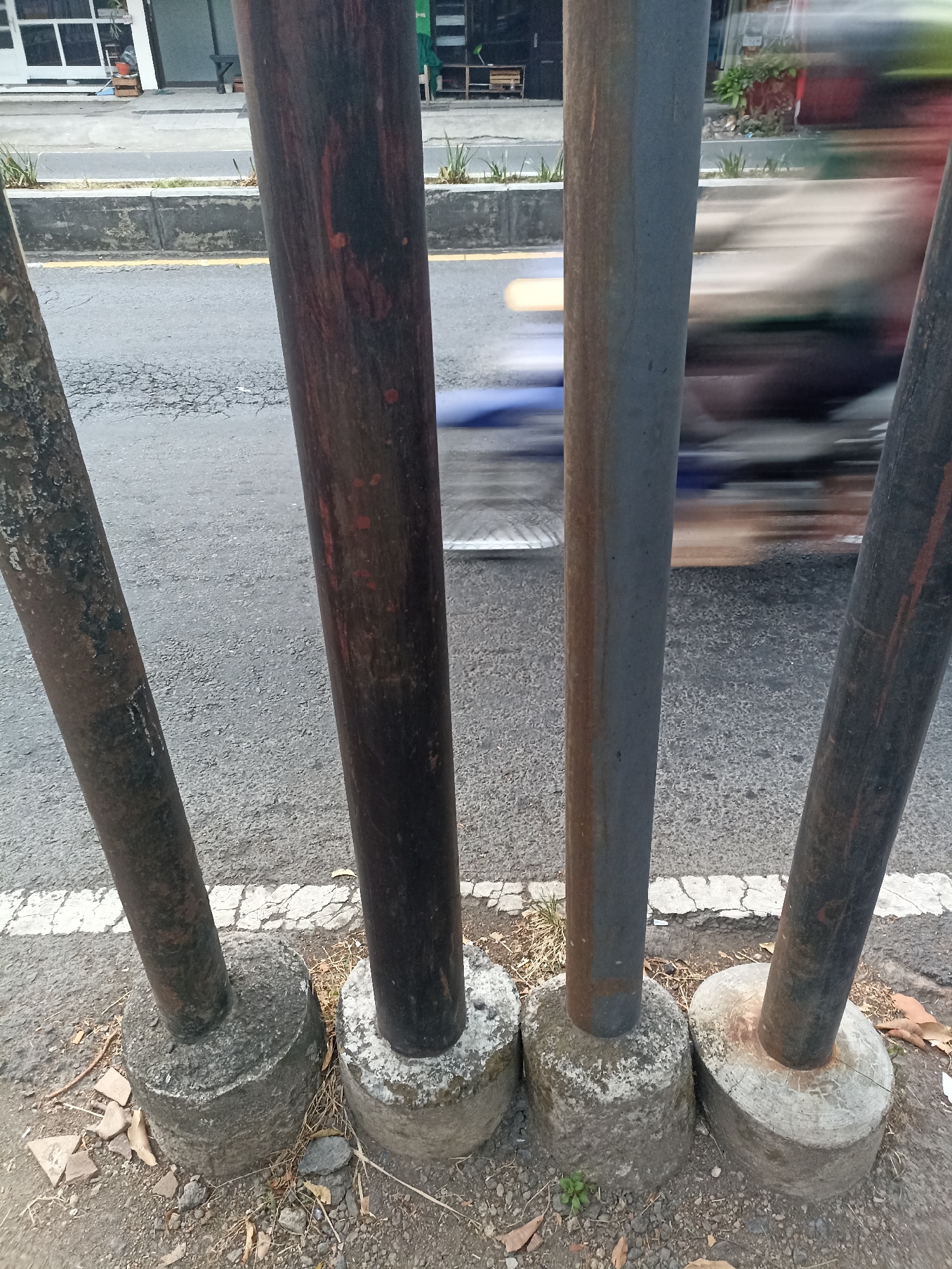 A low-angle, eye-level, medium shot captures four weathered metal poles standing vertically in the foreground, anchored to concrete bases on a roadside. The poles, varying in thickness and color, are positioned in a line and angled slightly towards the viewer. They are situated on a gravel and dirt surface with scattered dried leaves and debris. A cracked white line demarcates the edge of an asphalt road behind the poles, where a blurred vehicle, possibly a motorcycle, is in motion, indicated by streaks of light and color. In the background, a building with large windows and a shopfront is visible, suggesting an urban environment. The scene is likely located in Banyuwangi, Indonesia, given the style of the building and the presence of a busy street.
