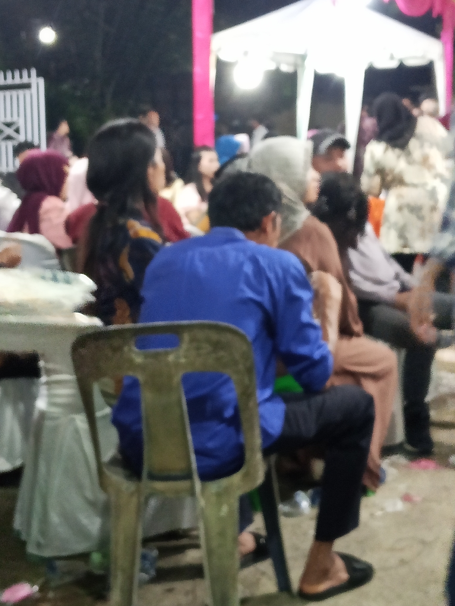 The image depicts an outdoor gathering, possibly a community event or celebration, in Perbarakan, Indonesia. In the foreground, a person wearing a bright blue long-sleeved shirt and dark pants is seated on a metal chair, facing away from the camera. Their left leg is extended, and they are wearing black flat shoes. To their left, another person in a muted red hijab and light-colored clothing is partially visible. In the background, several other individuals are present, some standing and others seated. A white tent is set up, suggesting a sheltered area for the event. The lighting appears to be artificial, with some light sources visible in the upper left quadrant of the image. The ground surface is indistinct but seems to be a rough or unpaved area. The overall atmosphere suggests a social occasion.