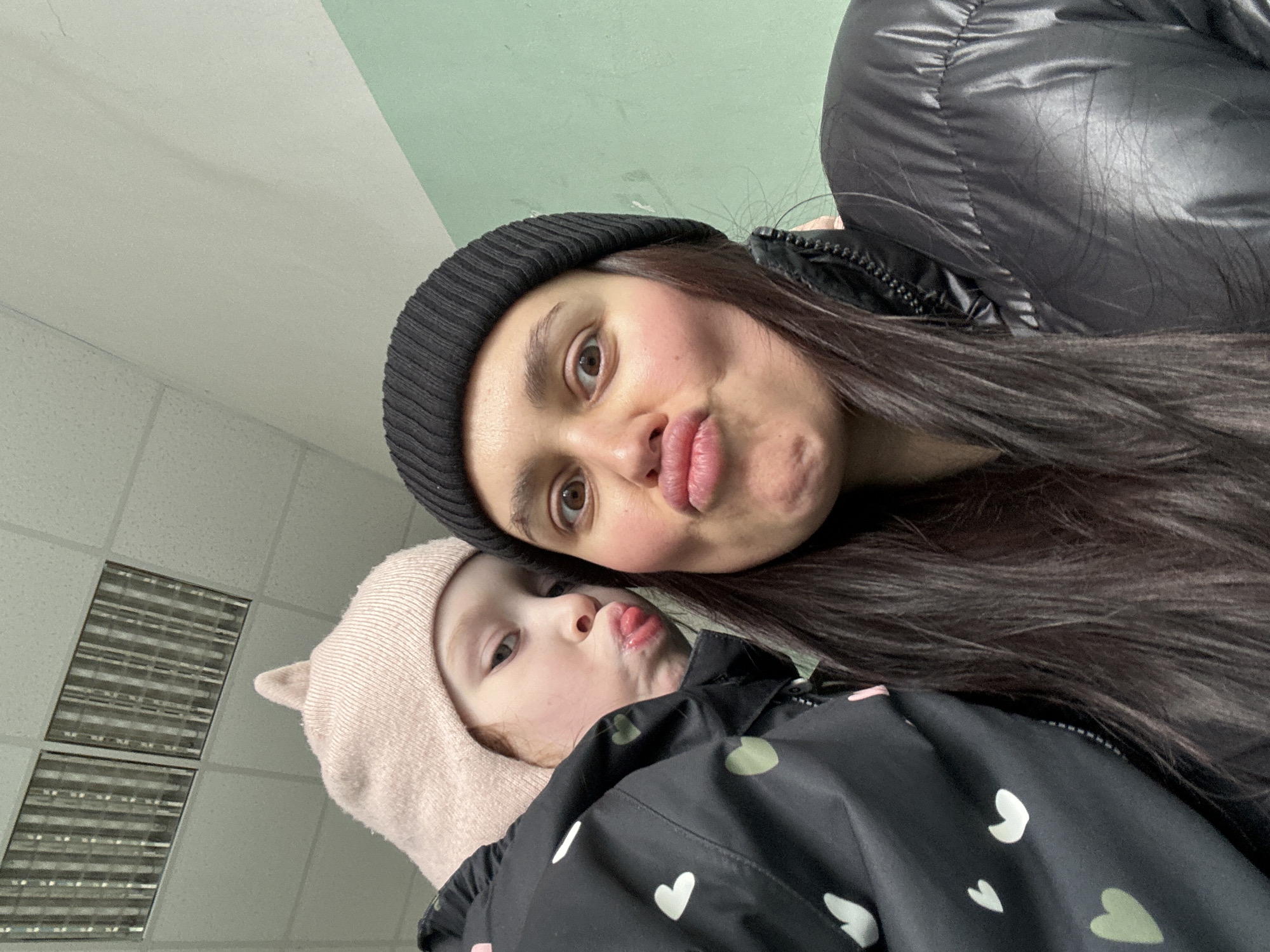 An adult woman and a young child are captured in a close-up, playful selfie, taken from a slightly upward angle. Both subjects are making puckered lip faces towards the camera.
The woman, on the right and slightly above the child, has long dark hair and is wearing a black ribbed beanie and a black puffy winter jacket. Her face shows rosy cheeks and a light brown eye is visible.
The child, positioned below and to the left of the woman, wears a light pink beanie with small ear details and a black jacket patterned with white and light green hearts. The child's tongue is slightly visible between the puckered lips.
The background consists primarily of a light-colored ceiling with acoustic tiles. Part of the ceiling transitions to a pale green color. In the upper left quadrant, two rectangular fluorescent light fixtures are visible, indicating an indoor setting.
The clothing suggests cold weather, appropriate for the location of Chaikovsky, Russia. The overall mood of the image is affectionate and lighthearted. No visible text is present.