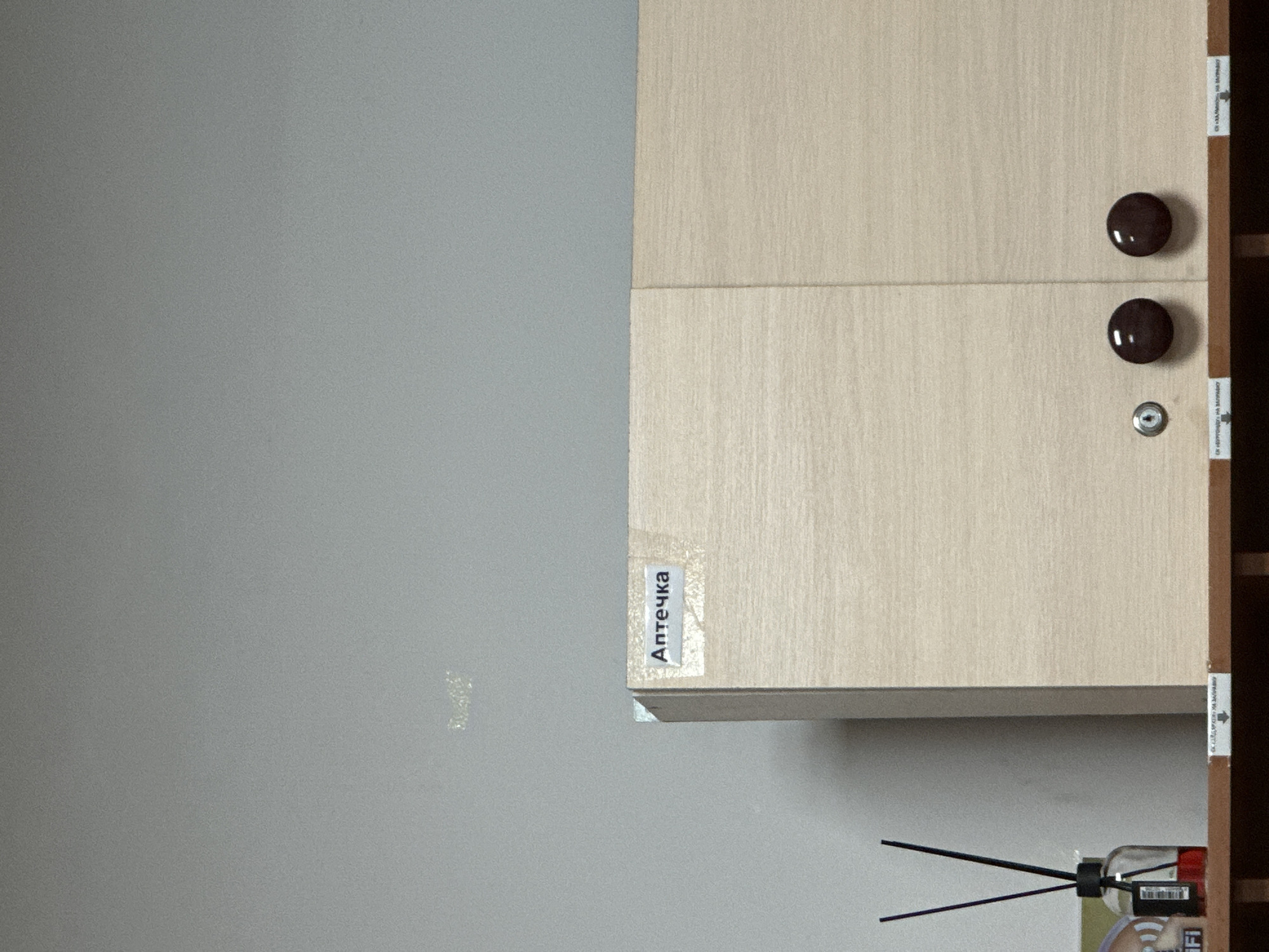 The image shows a close-up of a cabinet with a label that reads "Аптечка" (meaning "First Aid Kit" in Russian). The cabinet is light wood grain and has two dark brown circular handles. To the left of the cabinet, a bottle of red liquid with black reeds is visible, likely an air freshener. The background is a plain, light grey wall with some indistinct markings. The scene appears to be indoors. The image does not provide any specific information about the location in Kadamjay, Kyrgyzstan.