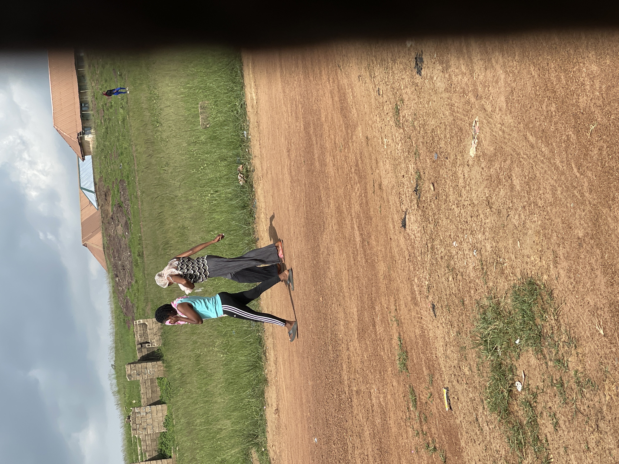 In Durumi, Nigeria, two young women walk side-by-side on a dirt road. The woman on the left wears a turquoise tank top and black leggings with white stripes. She has a pink cloth raised to her face. The woman on the right wears a black and white patterned sleeveless top and long, dark grey pants. She also has a light-colored cloth draped around her head and shoulders. They are walking past a grassy field with some scattered debris. In the background, there are partially constructed stone structures and buildings with orange roofs. The sky is partly cloudy.