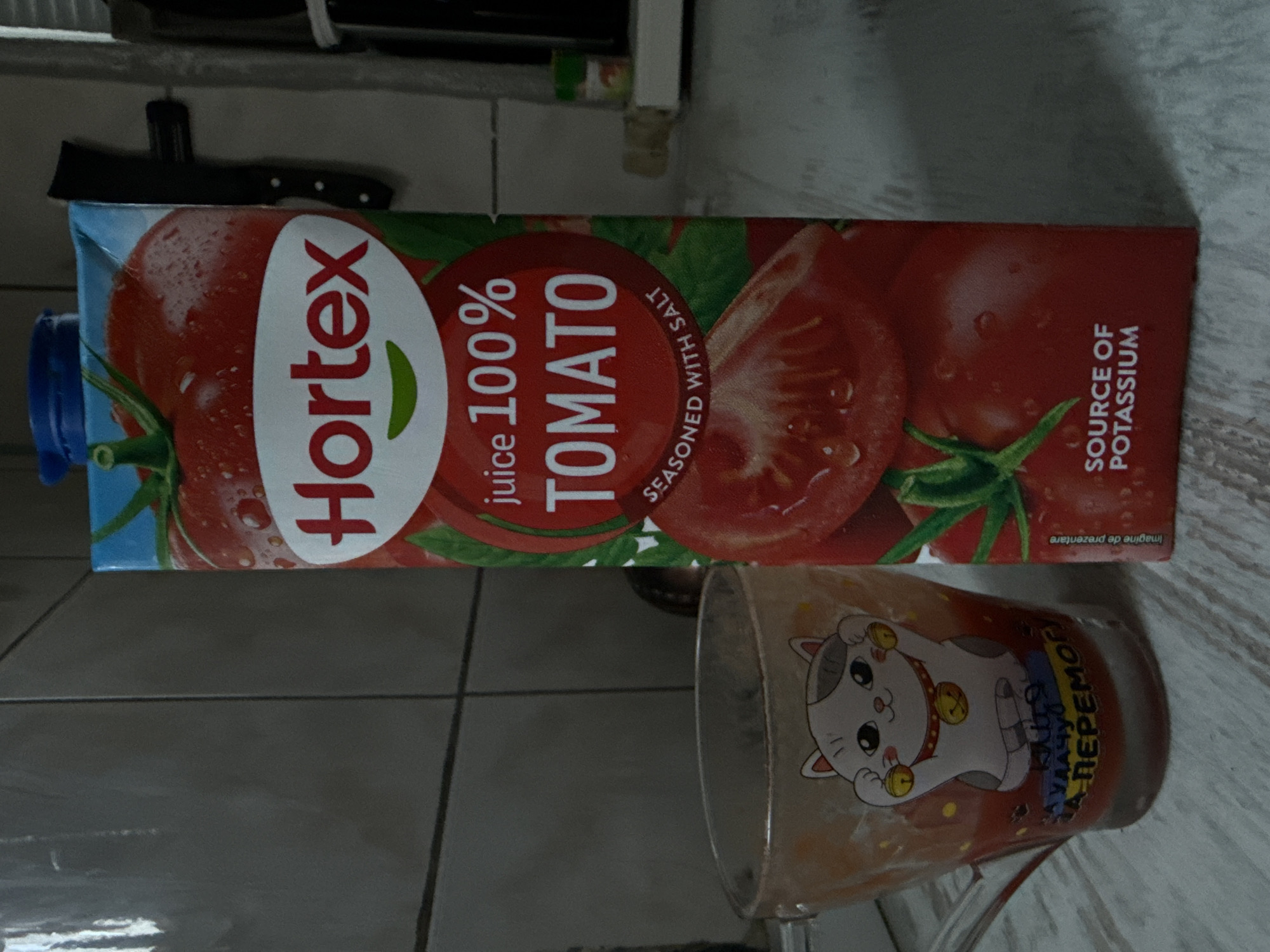 The scene, located in Uzhhorod, Ukraine, displays a Hortex brand 100% Tomato juice carton standing upright on a light-colored, subtly textured countertop. The carton is predominantly red with images of tomatoes and water droplets, featuring a blue screw cap. Visible text on the carton includes "Hortex," "juice 100% TOMATO," "SEASONED WITH SALT," and "SOURCE OF POTASSIUM." To the left of the carton, a clear glass mug containing a red liquid, consistent with tomato juice, is filled approximately one-third. The mug is decorated with a white Maneki-neko (lucky cat) graphic holding golden bells, and beneath it, Ukrainian text reads "Кіця на удачу За Перемогу!" (Kitty for luck, For Victory!). The background consists of a grey tiled wall with horizontal grout lines. In the far right background, a dark knife is mounted vertically, and a small container with a green cap rests on a white shelf.