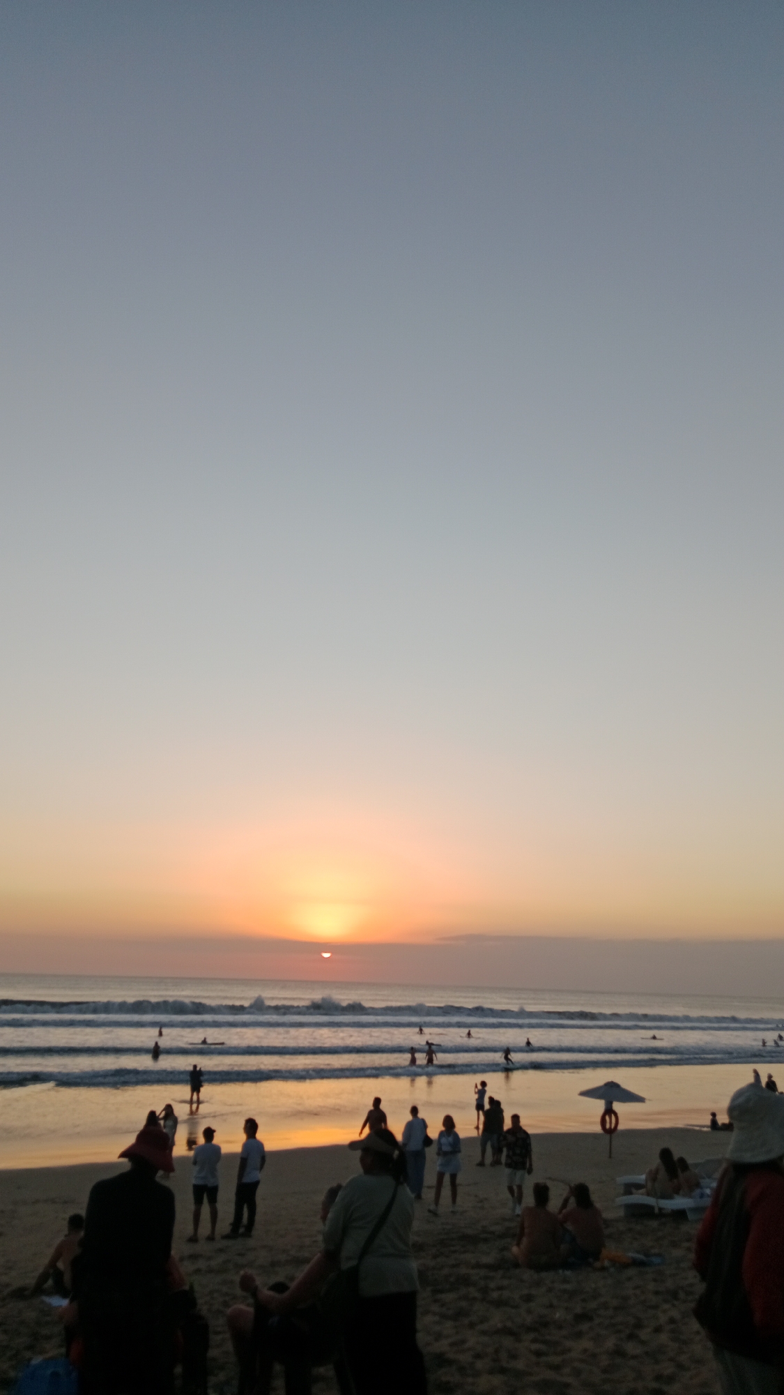 A vertical, low-angle shot captures a crowded beach scene at sunset in Kuta, Indonesia. The sky transitions from a pale blue at the top to a warm orange and yellow glow near the horizon, where the sun is a small, glowing orb partially obscured by clouds. Waves roll onto the shore, with several figures visible in the water, some appearing to be surfing. The sandy beach is populated with numerous people of various ages, dressed in casual attire. Some individuals are walking, others are standing or sitting, and a few are engaged in conversation. Among the beachgoers, there are umbrellas, lounge chairs, and a life preserver attached to a pole. The overall atmosphere suggests a lively and popular tourist destination.