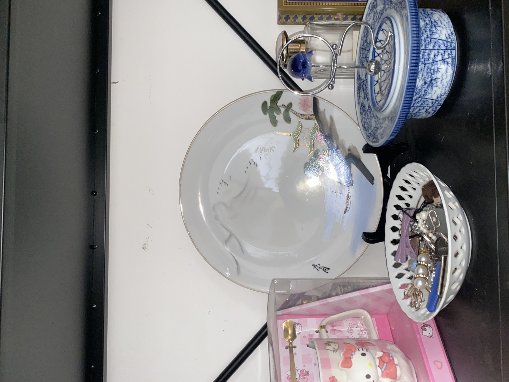 This is a medium shot, eye-level photograph of a shelf in Davao City, Philippines, displaying various decorative and collectible items. The shelf appears to be made of black material, possibly wood, and is mounted against a white wall.
Dominating the center of the frame is a decorative plate with a landscape design featuring mountains, trees, and birds. The plate rests on a black stand. To the left of the plate is a pink and white Hello Kitty mug in its original packaging, along with a golden spoon. Next to the Hello Kitty mug is a white, lattice-patterned bowl filled with various small items, including keys, hair clips, and jewelry.
To the right of the decorative plate is a tiered blue and white ceramic dish. A small, silver stand with a blue glass top is placed on this dish. Behind the ceramic dish, a clear glass perfume bottle is visible, and partially behind that, a framed item with a gold border and a patterned blue and gold design. The overall arrangement suggests a display of personal belongings or collectibles.