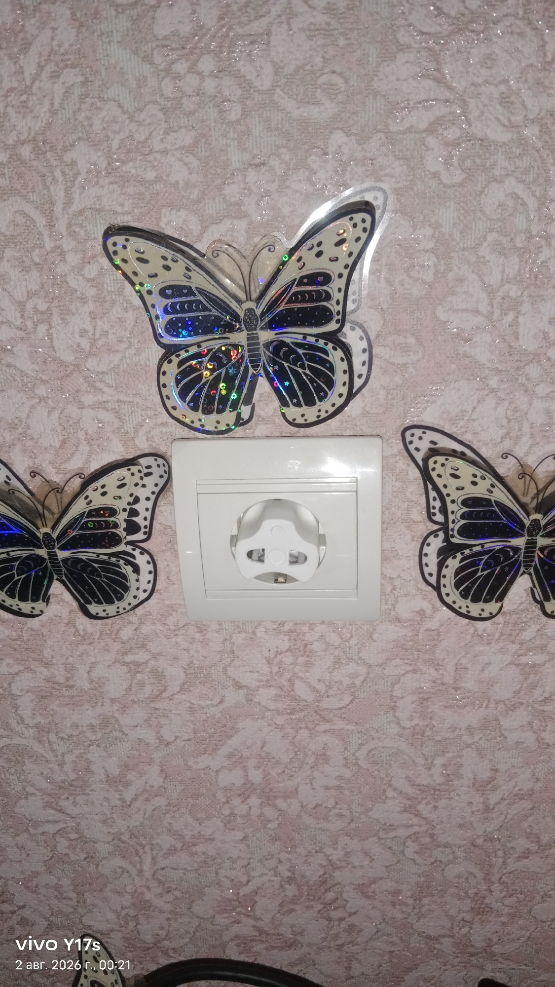 The image shows a white electrical outlet with a plastic child safety cover on a wall. Three decorative butterfly stickers are placed around the outlet: one above and two to the sides. The wall has a textured, pale pink wallpaper with a subtle damask pattern. The lighting suggests an indoor setting. The bottom left corner displays a "vivo Y17s" watermark and the date "2 авг. 2026 г., 00:21". There are no people visible in the image. The location is not explicitly stated to be in Rudny, Kazakhstan; this information is external to the image content.